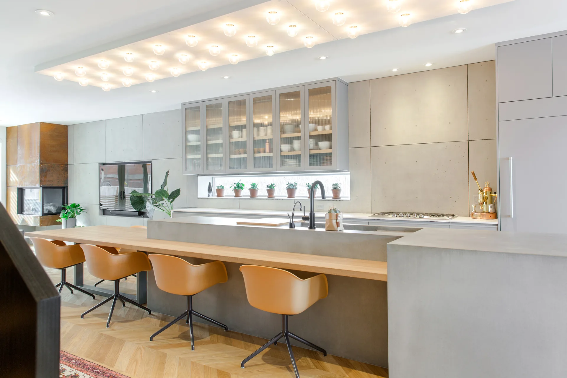 A modern kitchen featuring a concrete island with a wooden breakfast bar, tan swivel stools, and a grid of ceiling lights over herringbone floors.