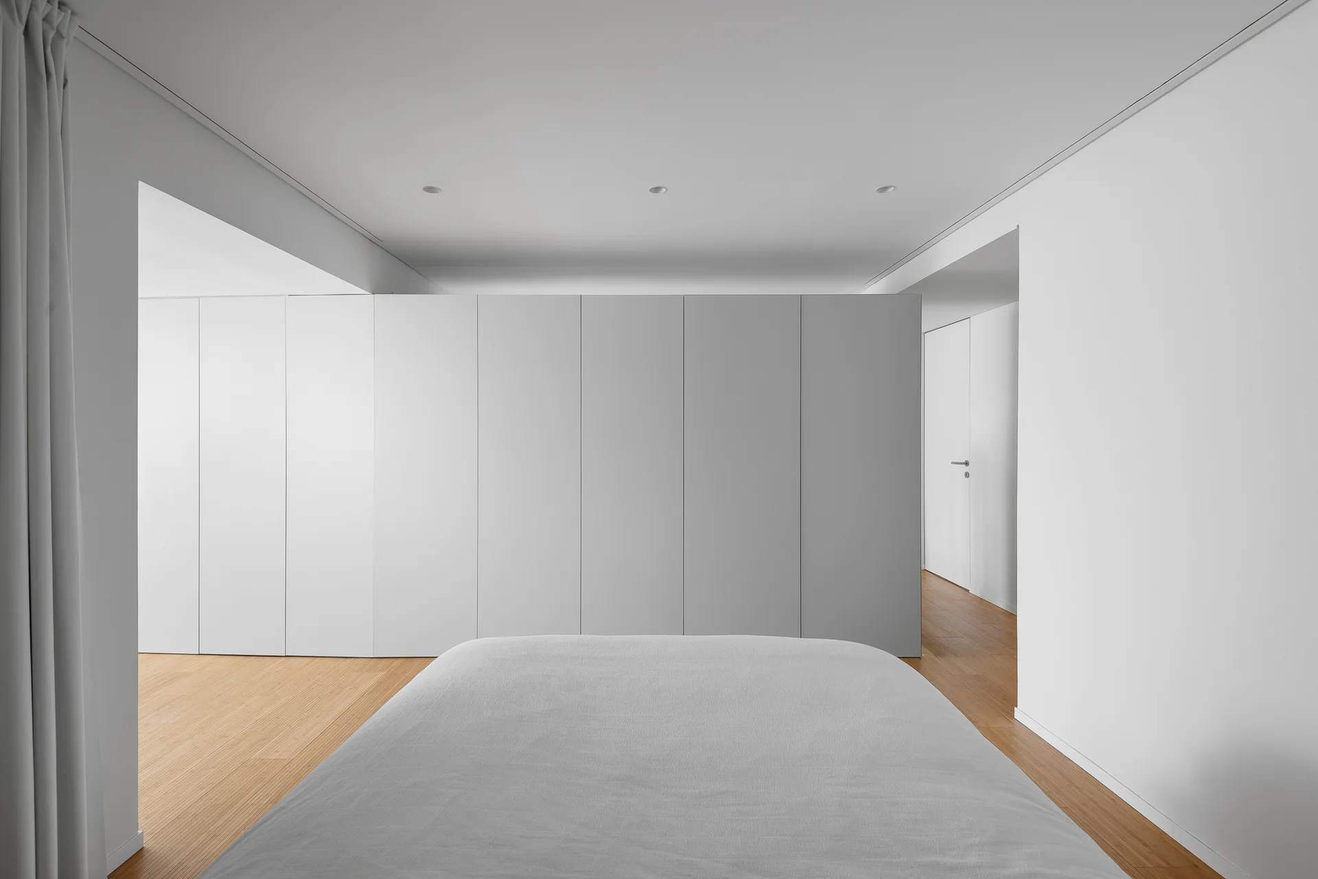 A minimalist bedroom features a grey bed in the foreground, a large seamless white wardrobe wall, light wood flooring, and recessed ceiling lights.