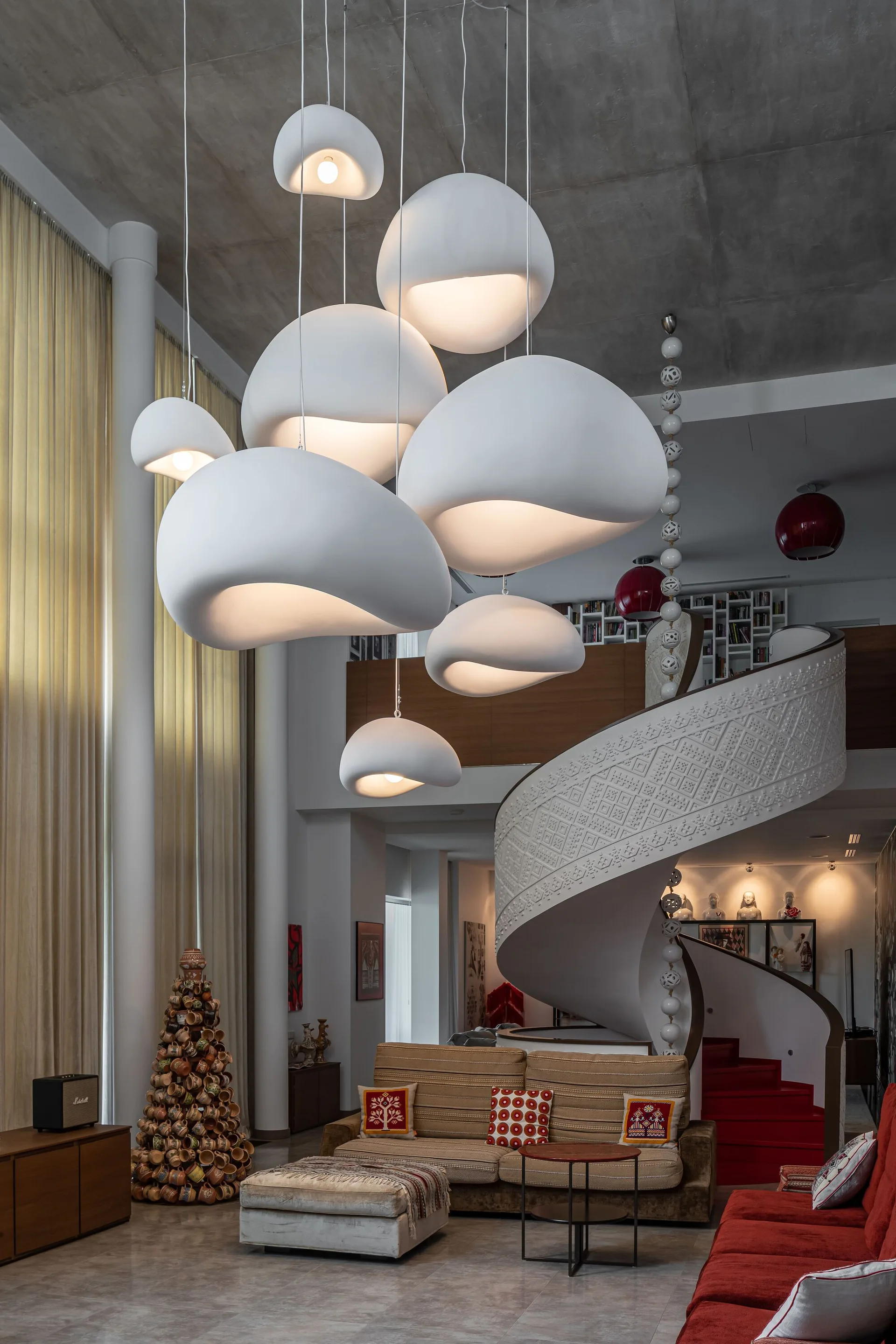 A spacious living room features a cluster of white organic pendant lights hanging below a concrete ceiling near a carved white spiral staircase.