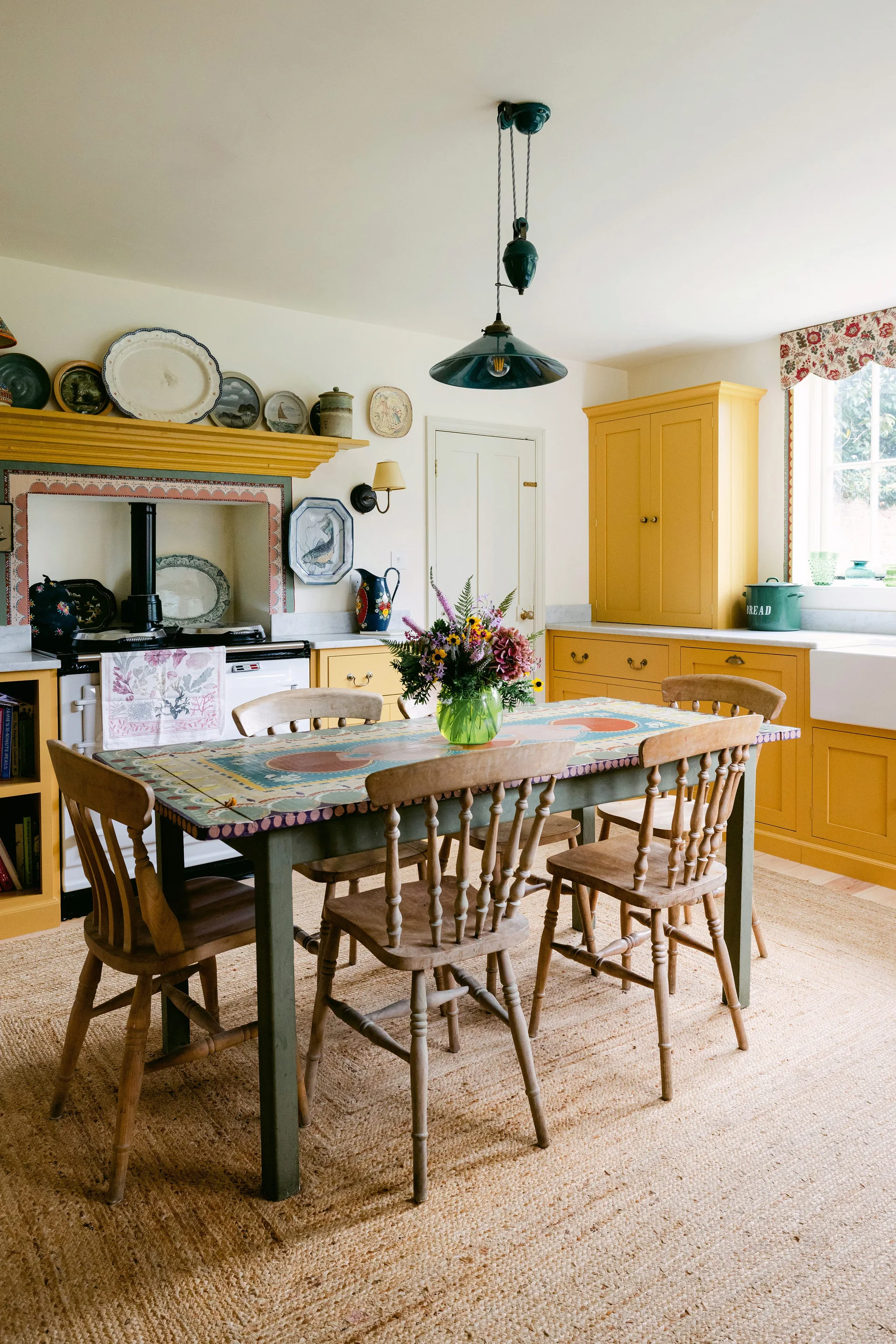A cottage kitchen features bright yellow cabinets, a patterned wooden dining table with chairs, and a natural fiber rug.