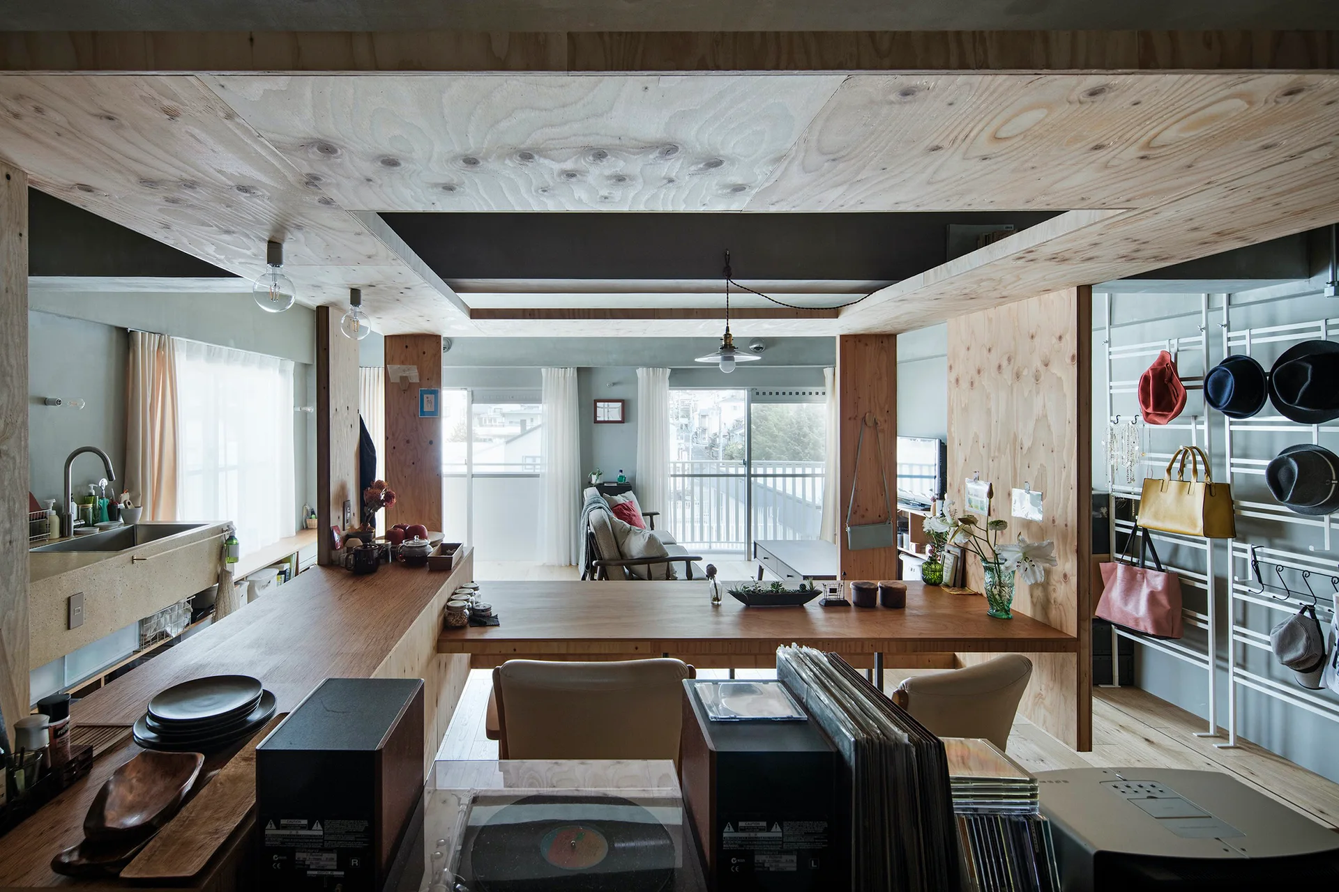 An open-concept home interior featuring a light wood kitchen counter, exposed plywood ceiling panels, and a living area in the background.