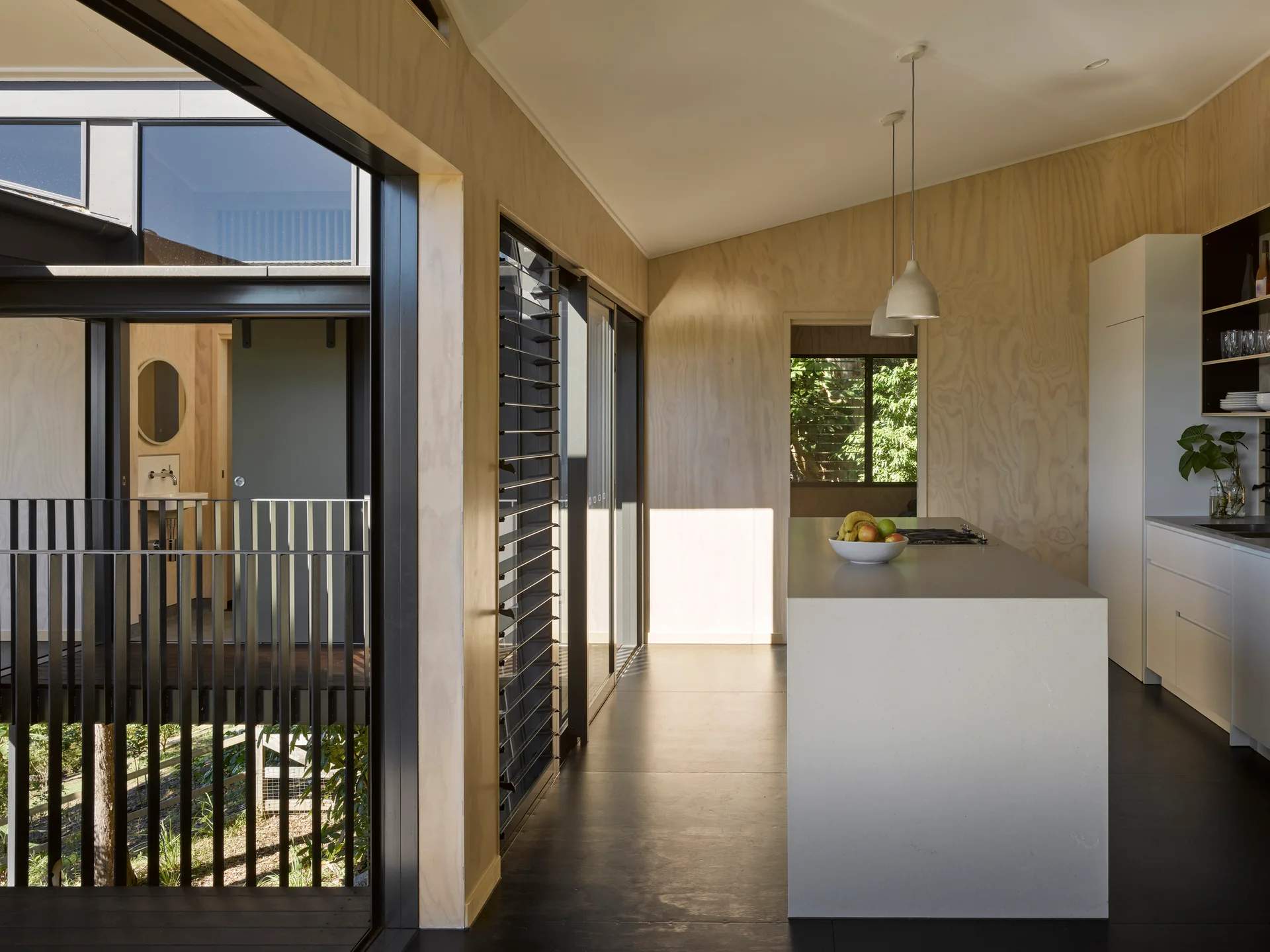 A minimalist kitchen featuring light plywood walls, a large solid grey island, white cabinets, dark floors, and louvered glass windows.