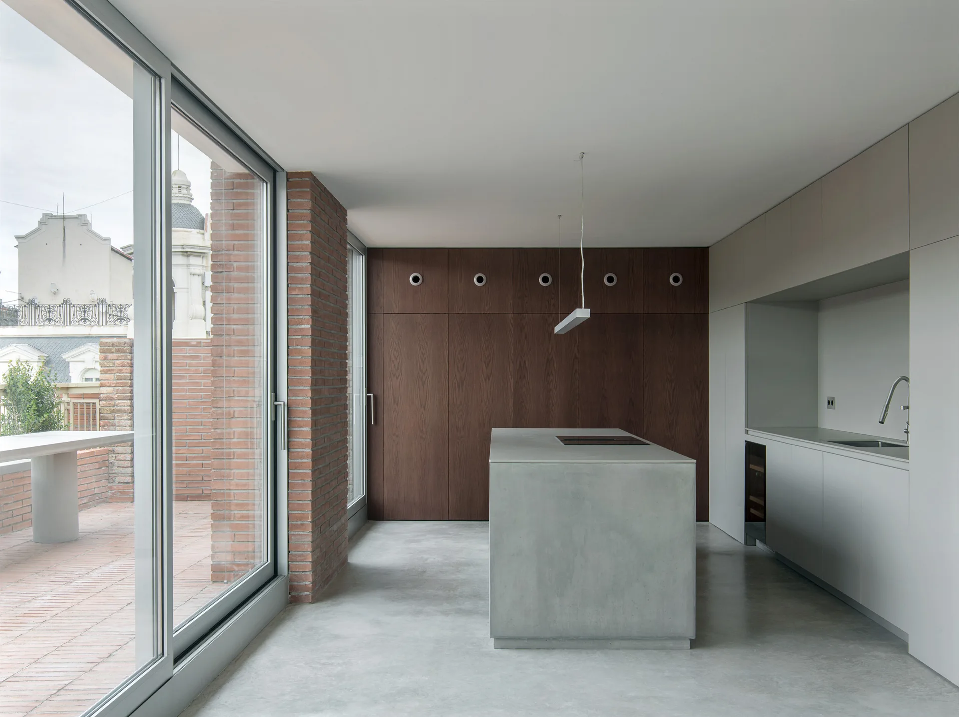A minimalist kitchen features a grey concrete island, dark wood storage cabinets, and sliding glass doors that open to a brick terrace.