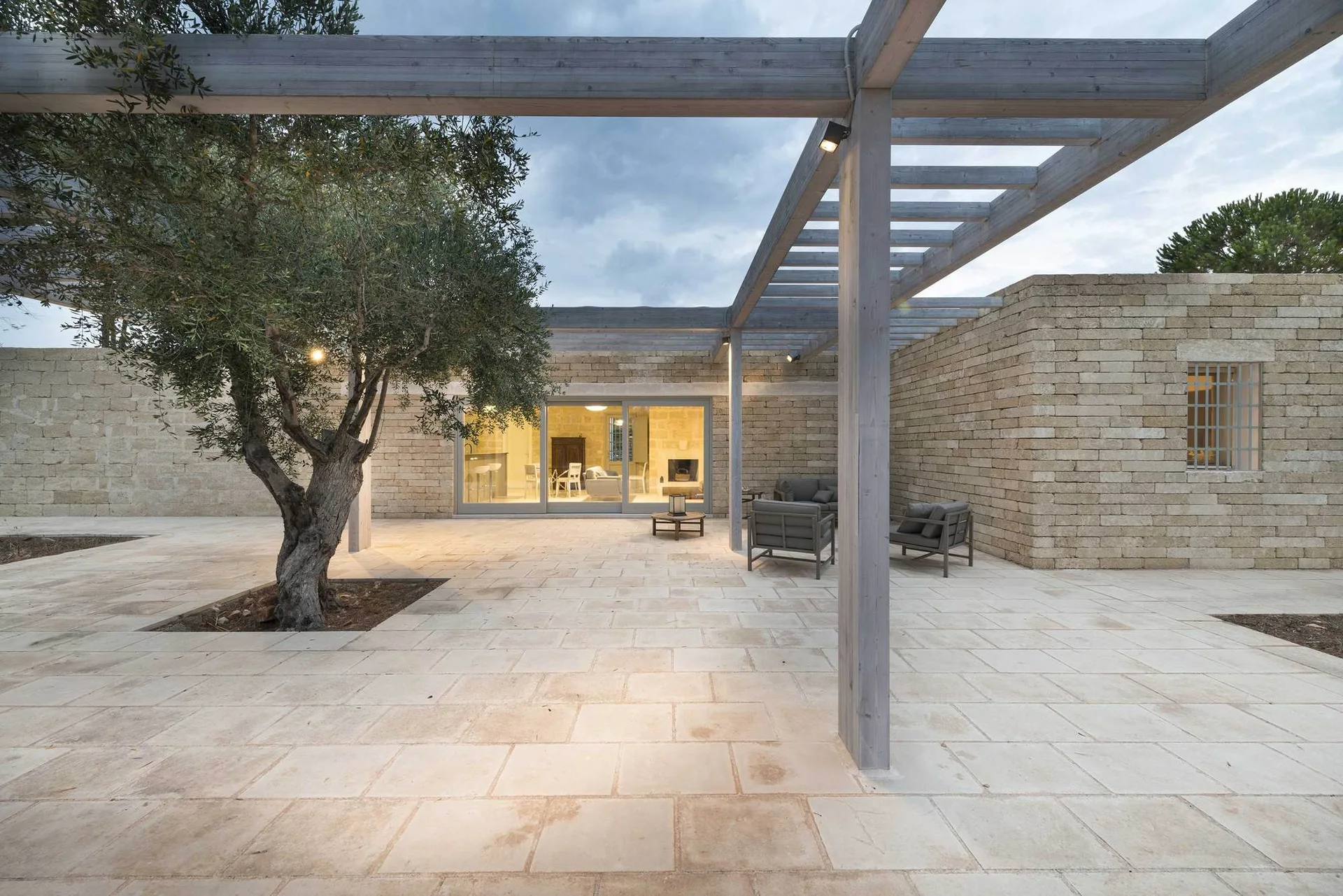 A paved stone patio features a weathered grey timber pergola, stacked stone walls, modern seating, and a mature olive tree.