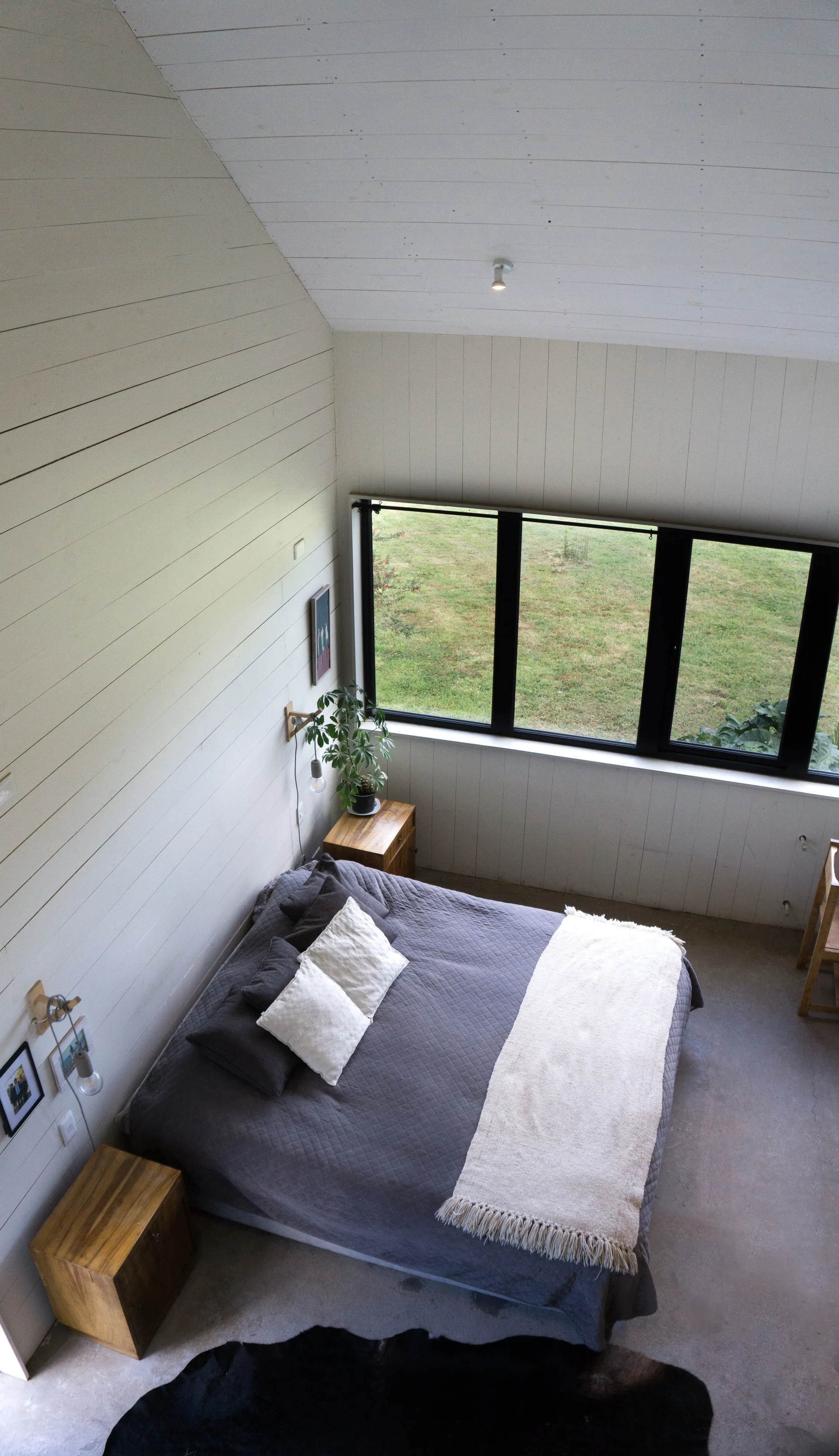 High-angle view of a minimalist bedroom with a grey bed, wooden nightstands, horizontal paneled walls, and a large window.