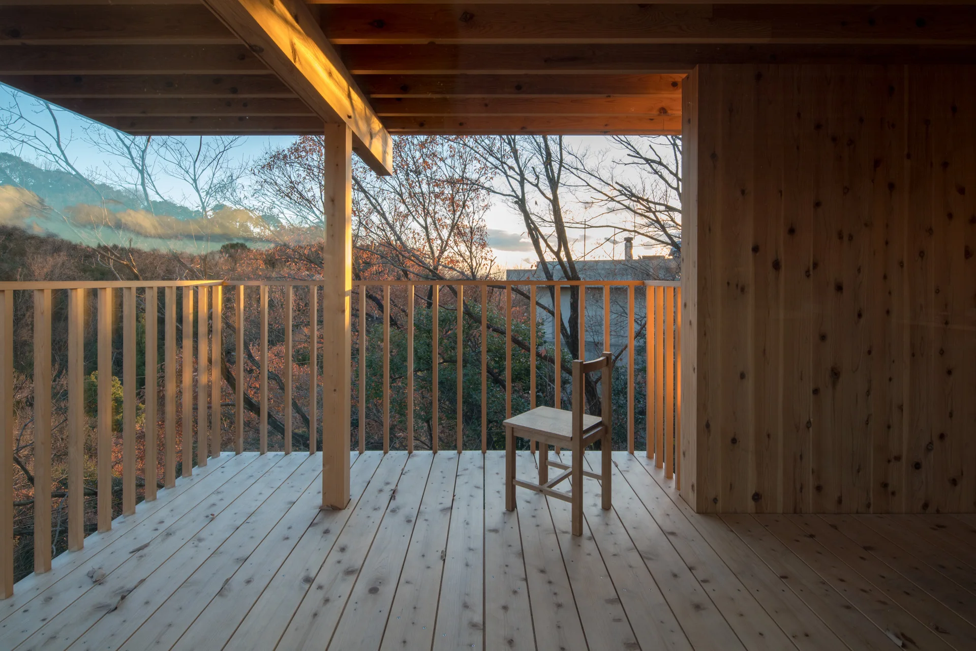 A minimalist wooden chair sits on a covered balcony built with natural timber planks and a vertical slat railing overlooking trees.