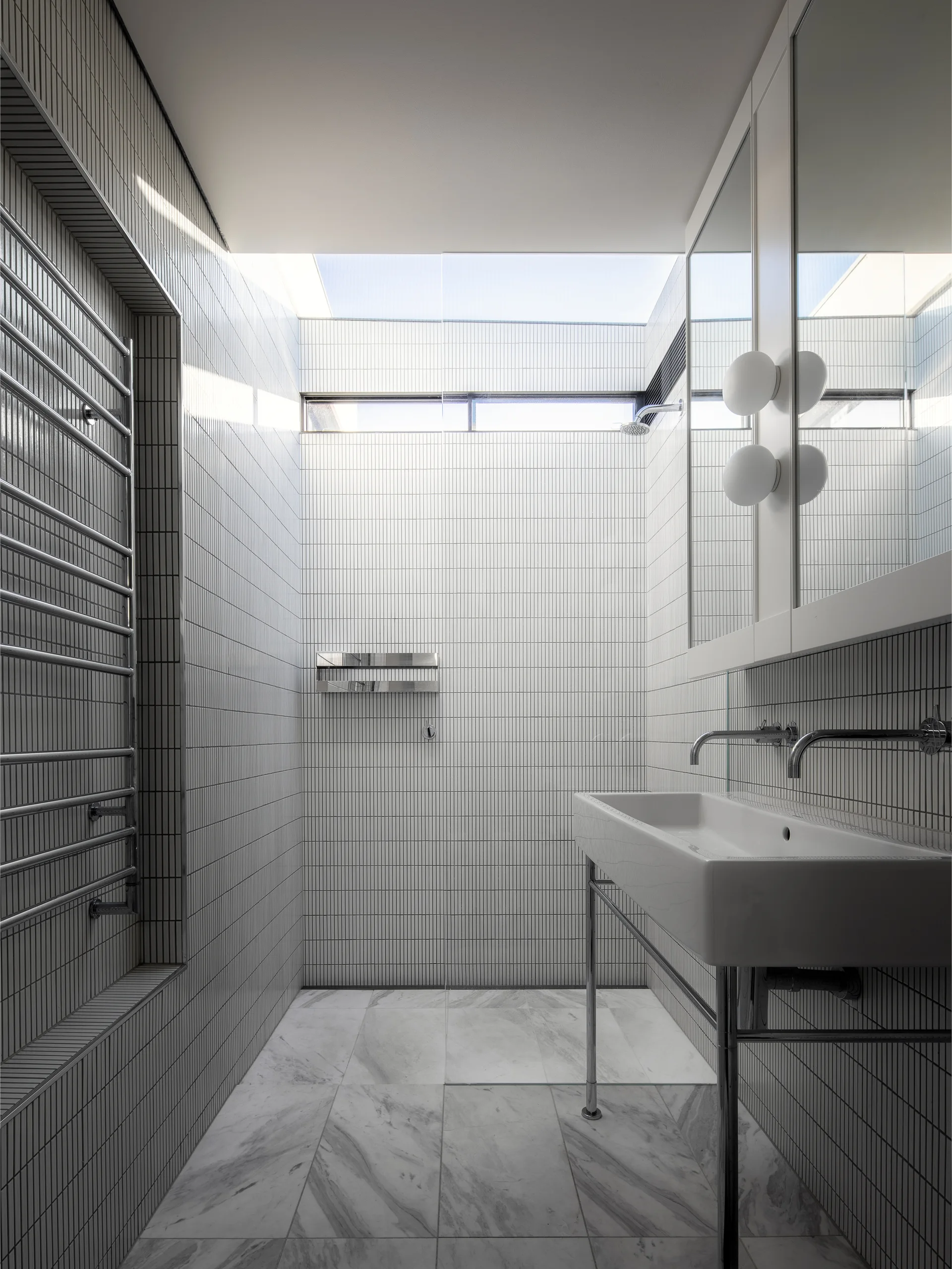 A narrow modern bathroom featuring vertical white wall tiles, a skylight, a console sink, and marble floor tiles.