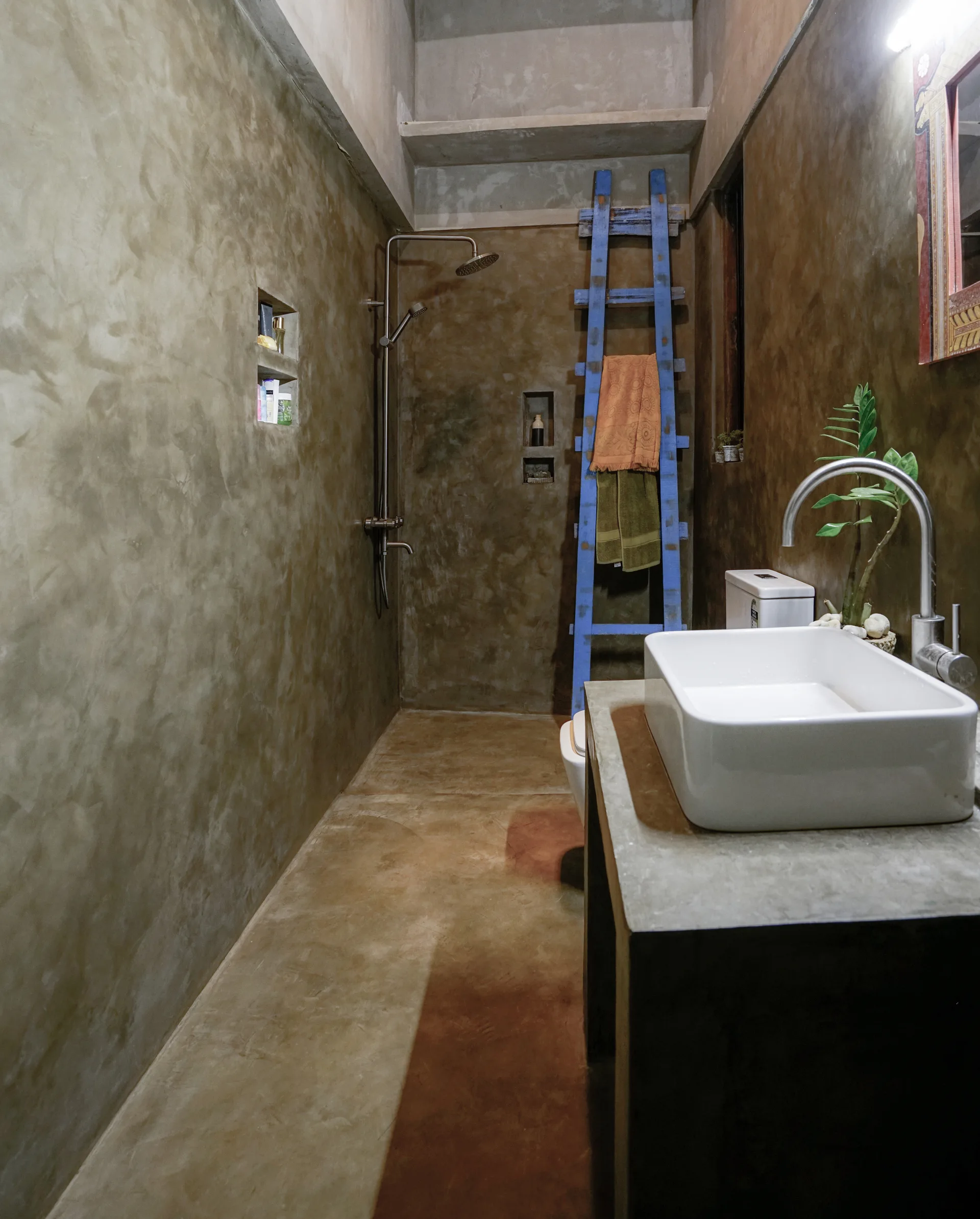 An industrial bathroom features textured concrete walls, a white ceramic vessel sink on a vanity, and a rustic blue wooden ladder holding towels.