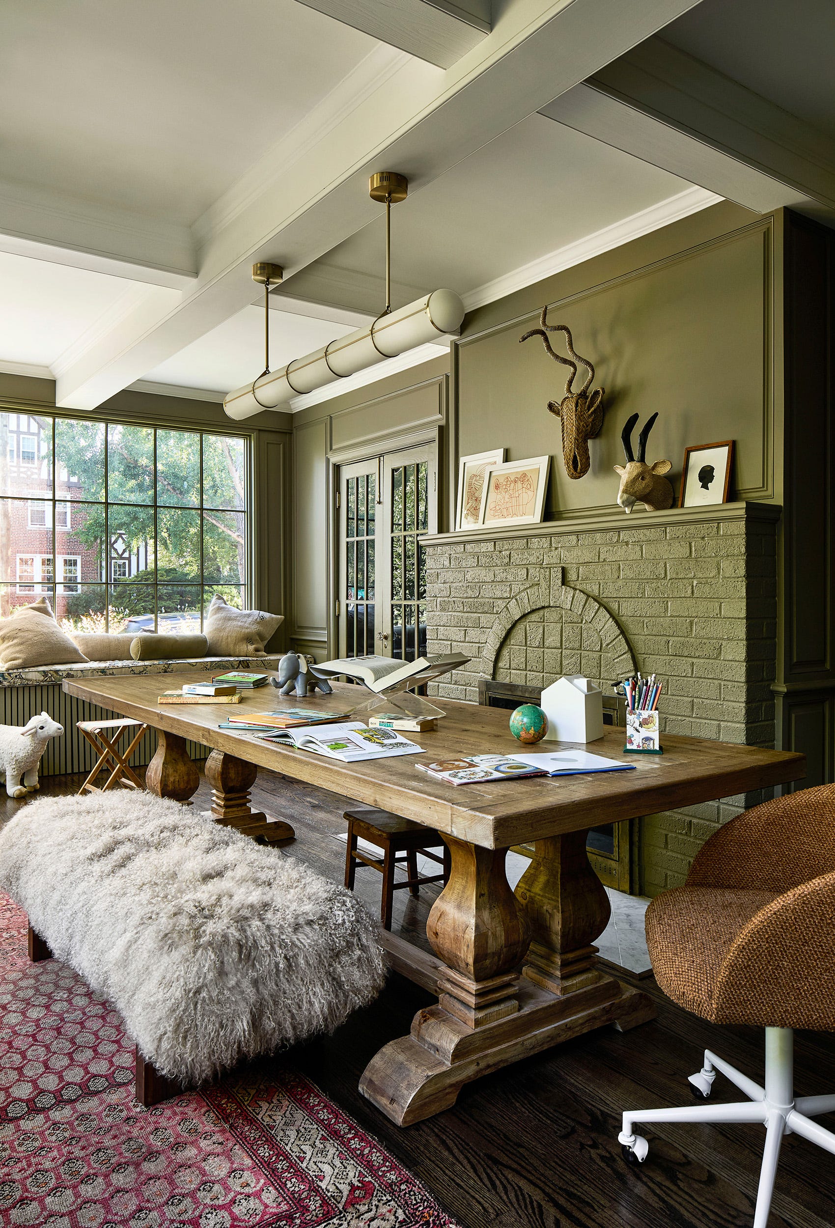 A playroom features a large rustic wooden table, a shaggy bench, and olive green paneled walls with a matching painted brick fireplace.