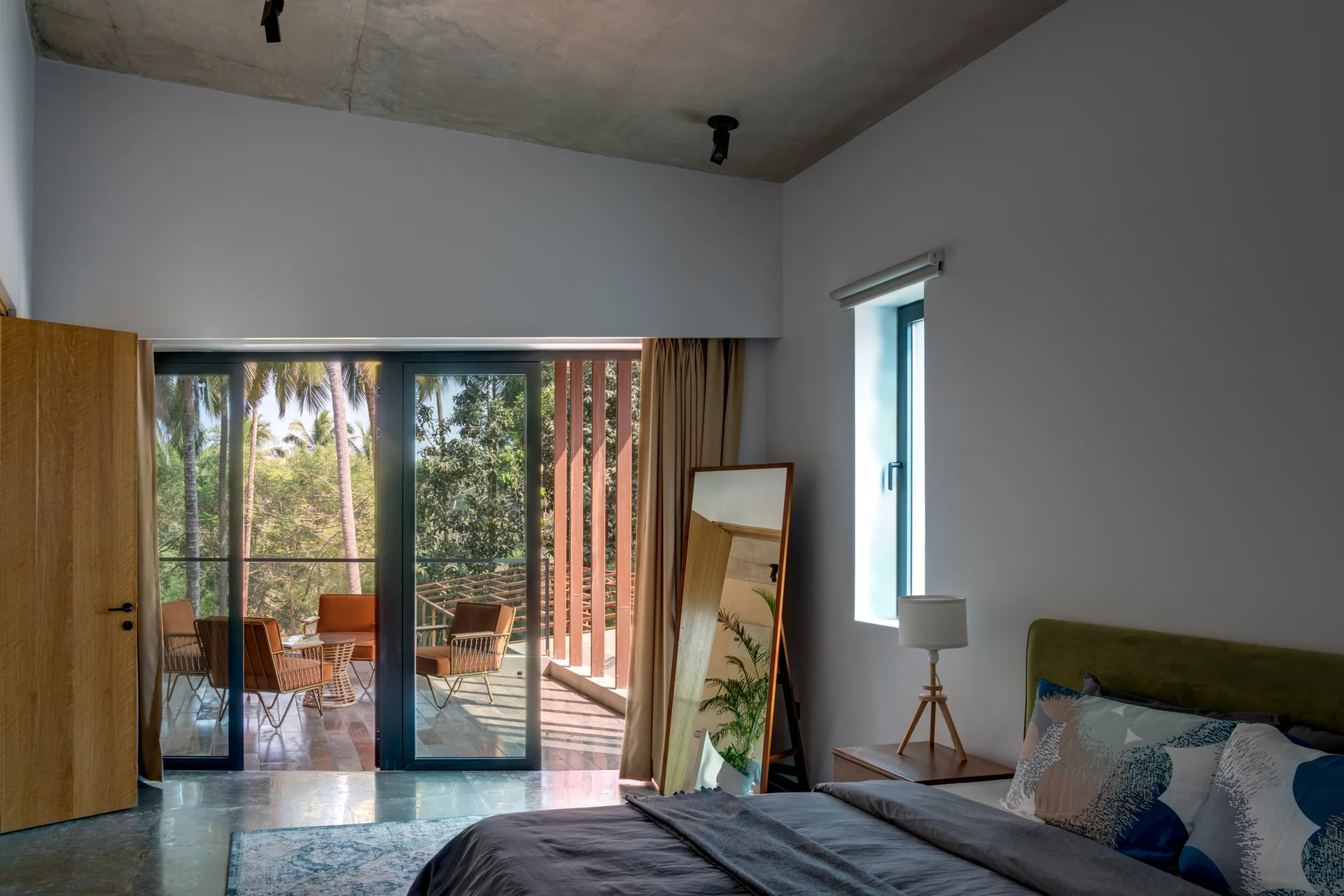 A modern bedroom features an exposed concrete ceiling, a bed with grey linens, a tall floor mirror, and sliding glass doors opening to a balcony.
