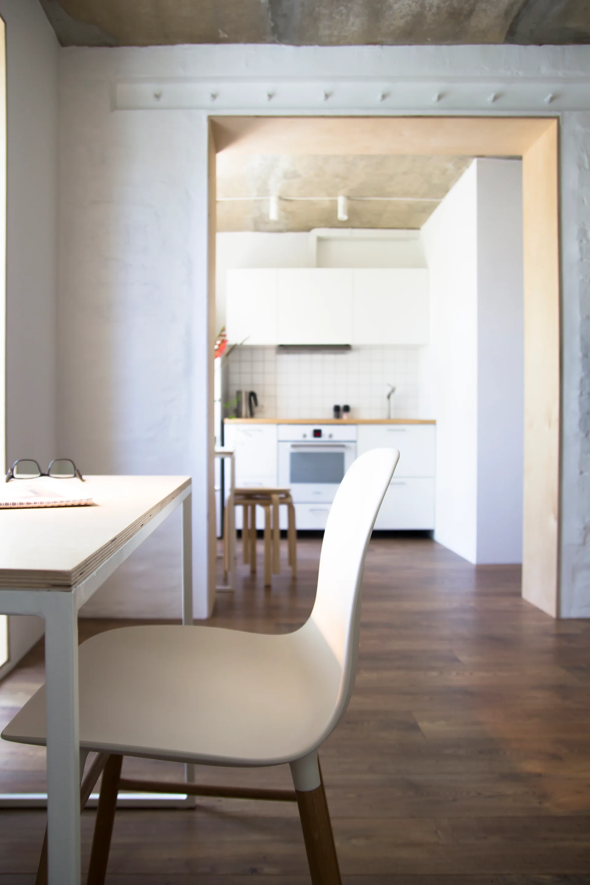 A white dining chair sits by a light wood desk, looking through a wood-lined doorway into a white kitchen with an exposed concrete ceiling.