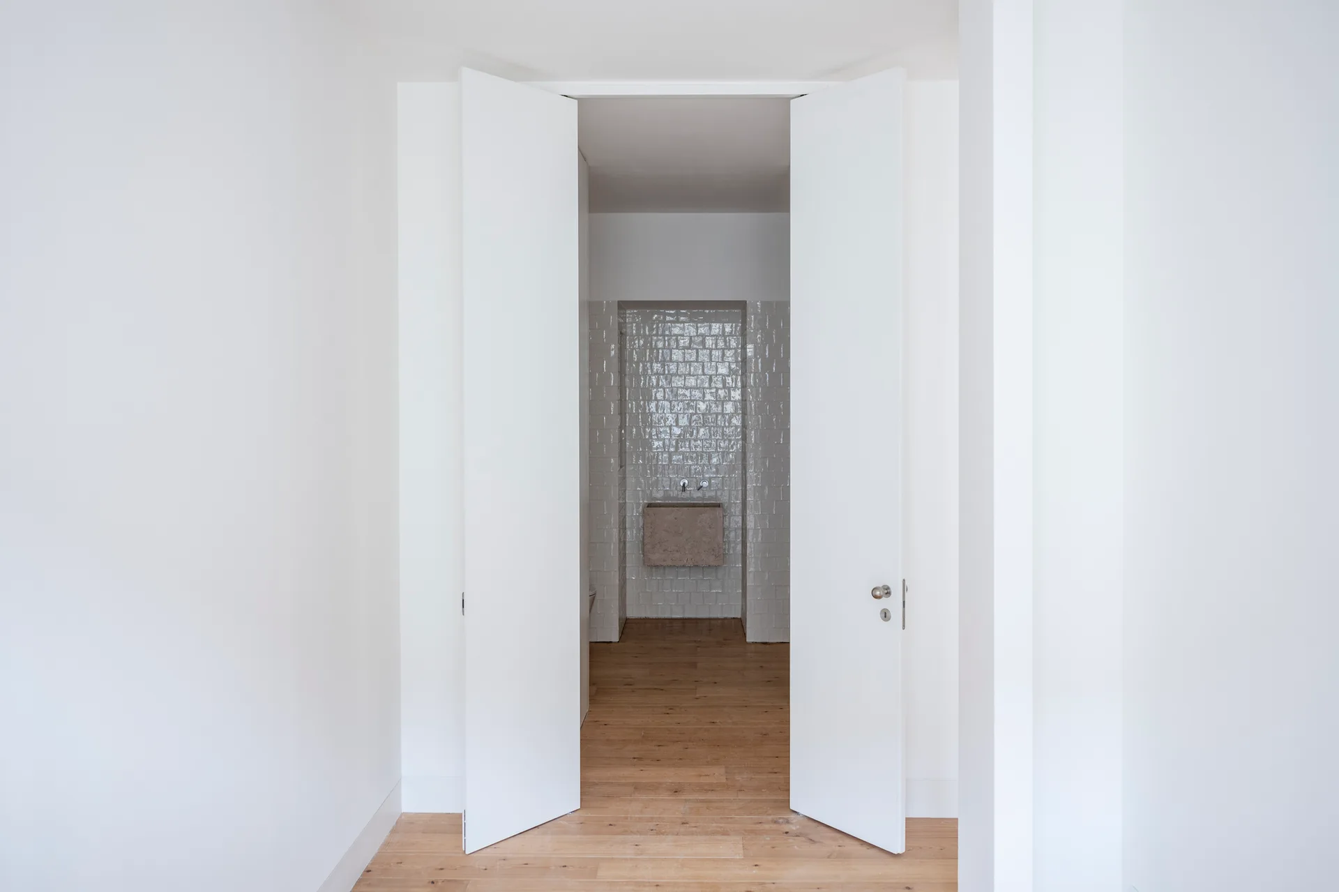 A view through open white double doors into a minimalist bathroom featuring a wall-mounted stone sink and glossy tiled walls.