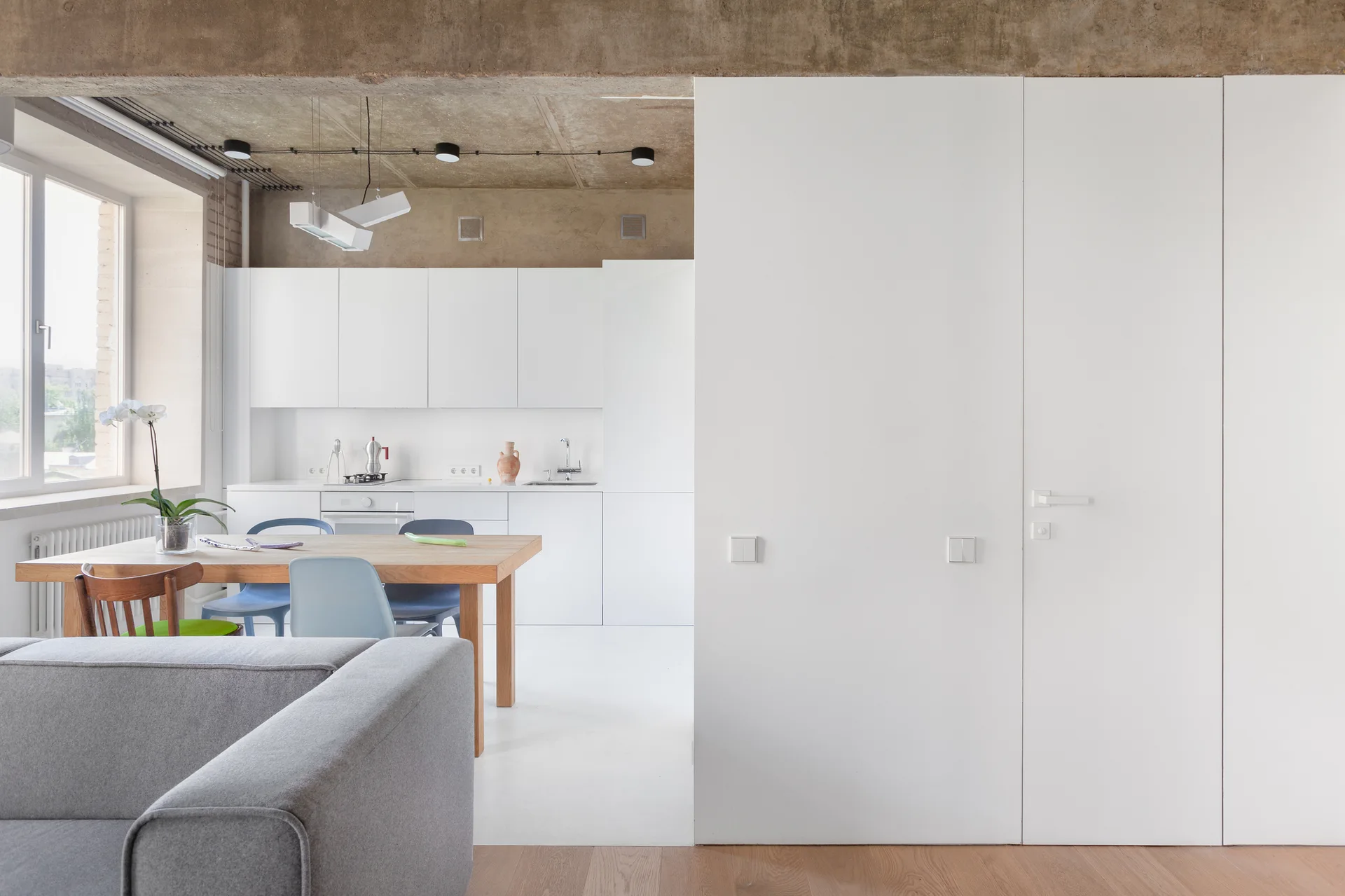 A modern open-plan kitchen features white flat-front cabinets, an exposed concrete ceiling, and a wooden dining table with chairs.