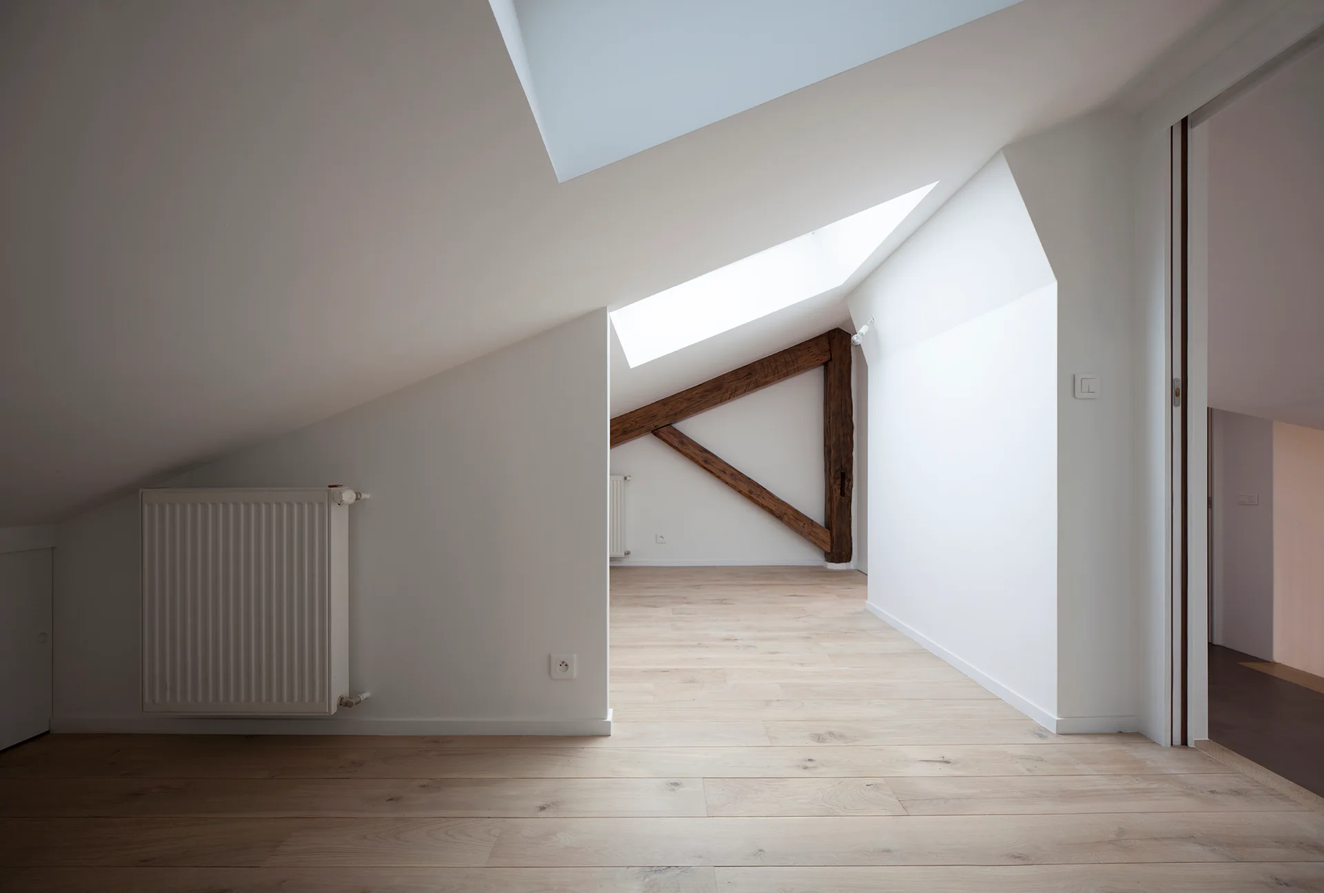 An empty attic room featuring white slanted walls, natural wood flooring, skylights, and a rustic wooden beam support.