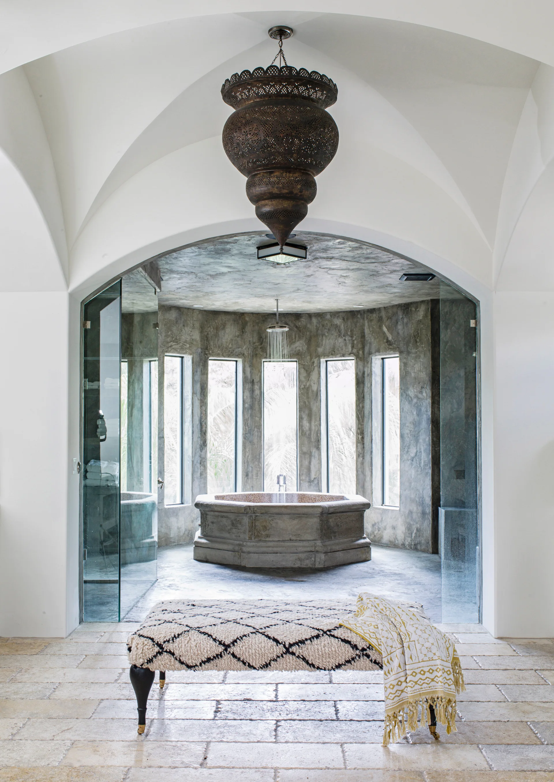 An arched entryway leads to a grey stone wet room featuring a central hexagonal bathtub, tall narrow windows, and a large ornate hanging metal pendant.