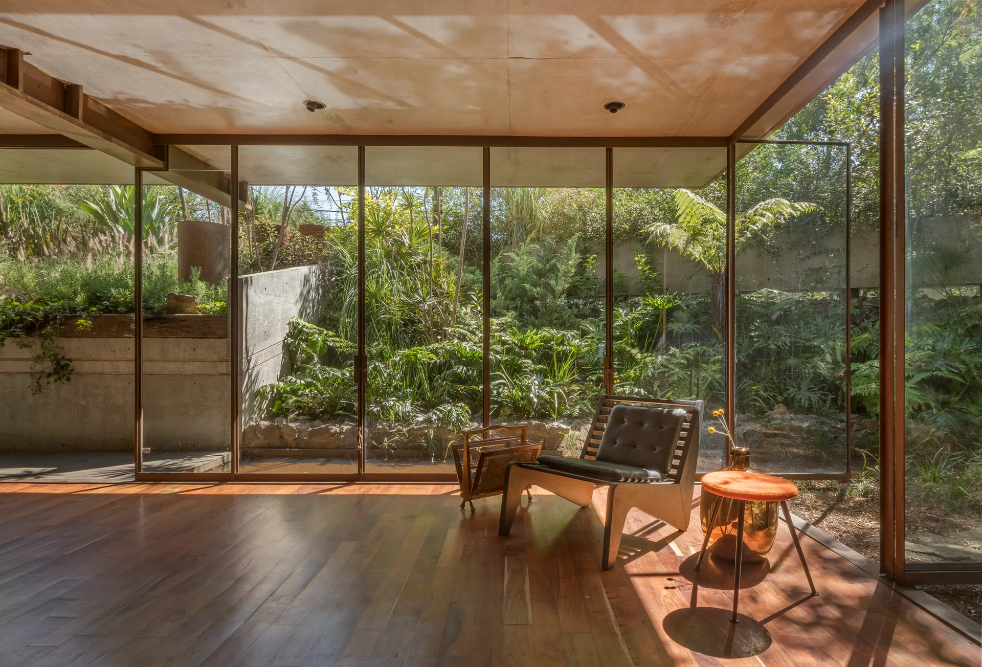 A minimalist room features a black leather-like lounge chair and an orange side table on hardwood floors next to a floor-to-ceiling glass wall.