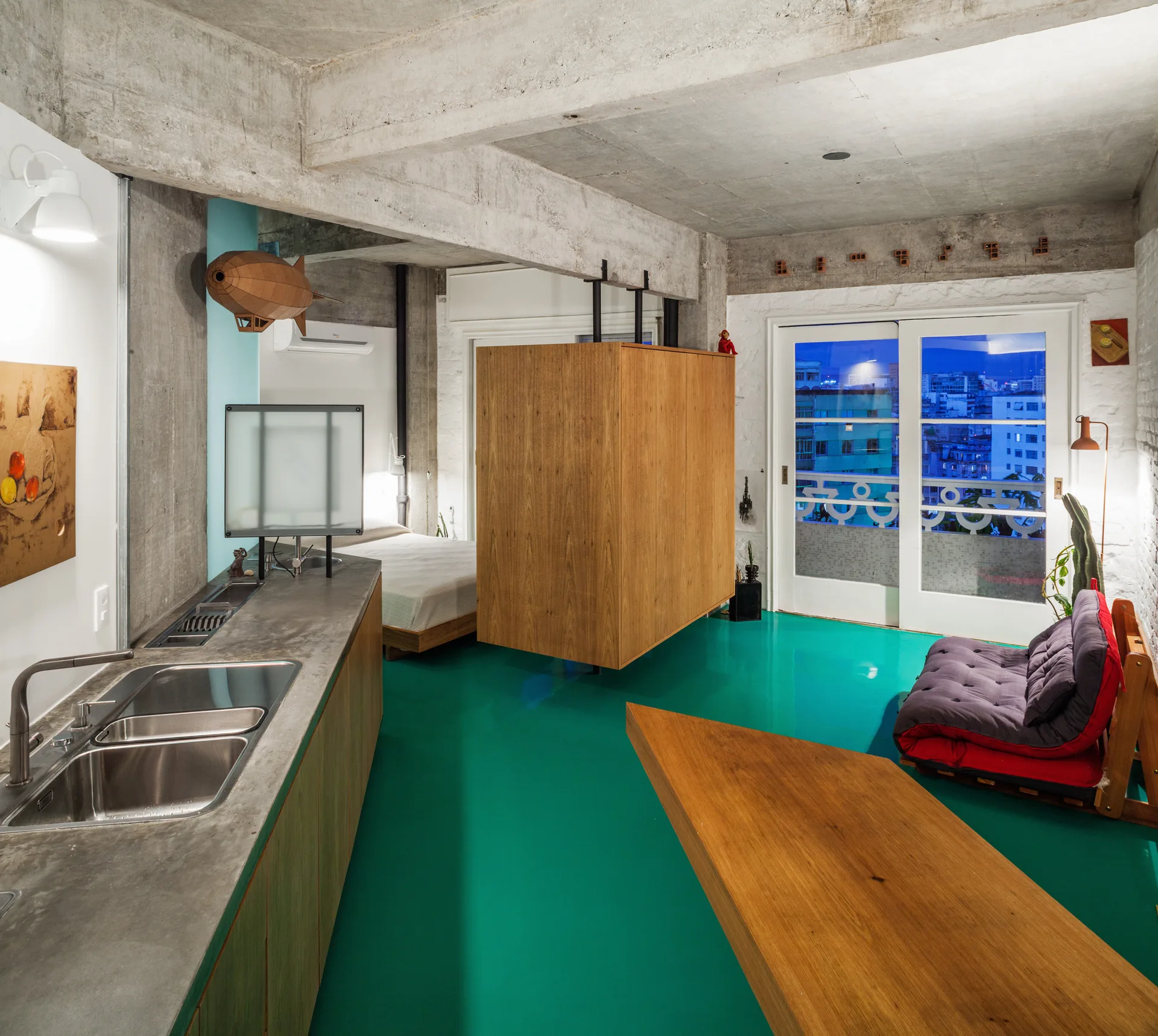 A modern studio apartment features a glossy teal floor, raw concrete ceiling beams, a central wooden room divider, and a concrete kitchen counter.