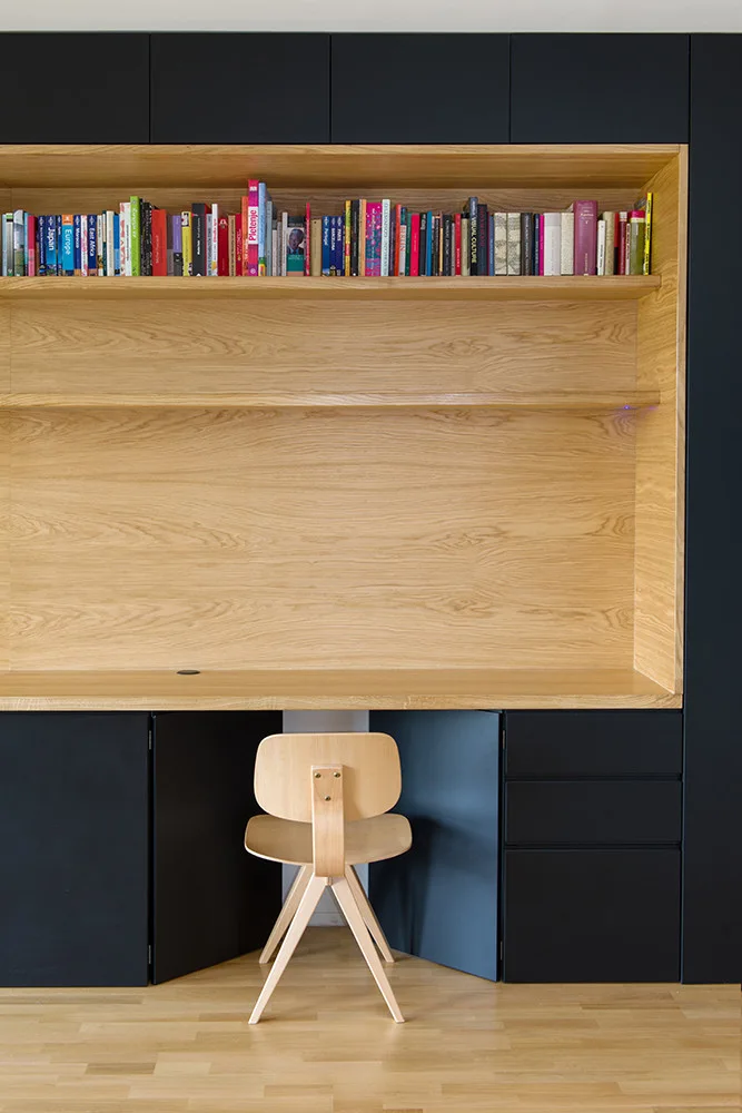 A modern home office featuring a built-in light wood desk and shelving unit recessed into a large matte black cabinetry wall with a wooden chair.