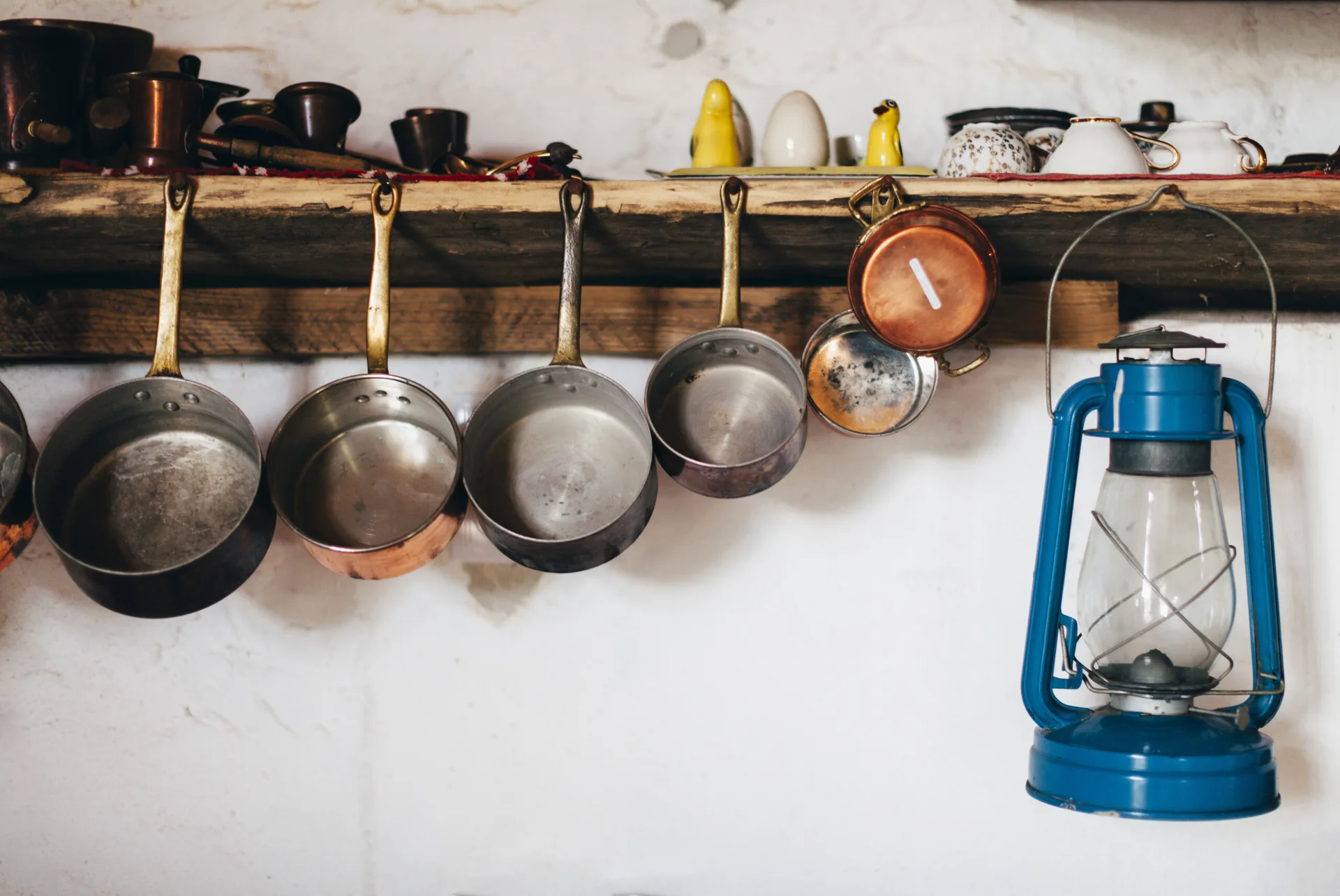 A row of copper and metal pots hangs underneath a rustic wooden shelf holding small ceramic items next to a blue oil lantern.
