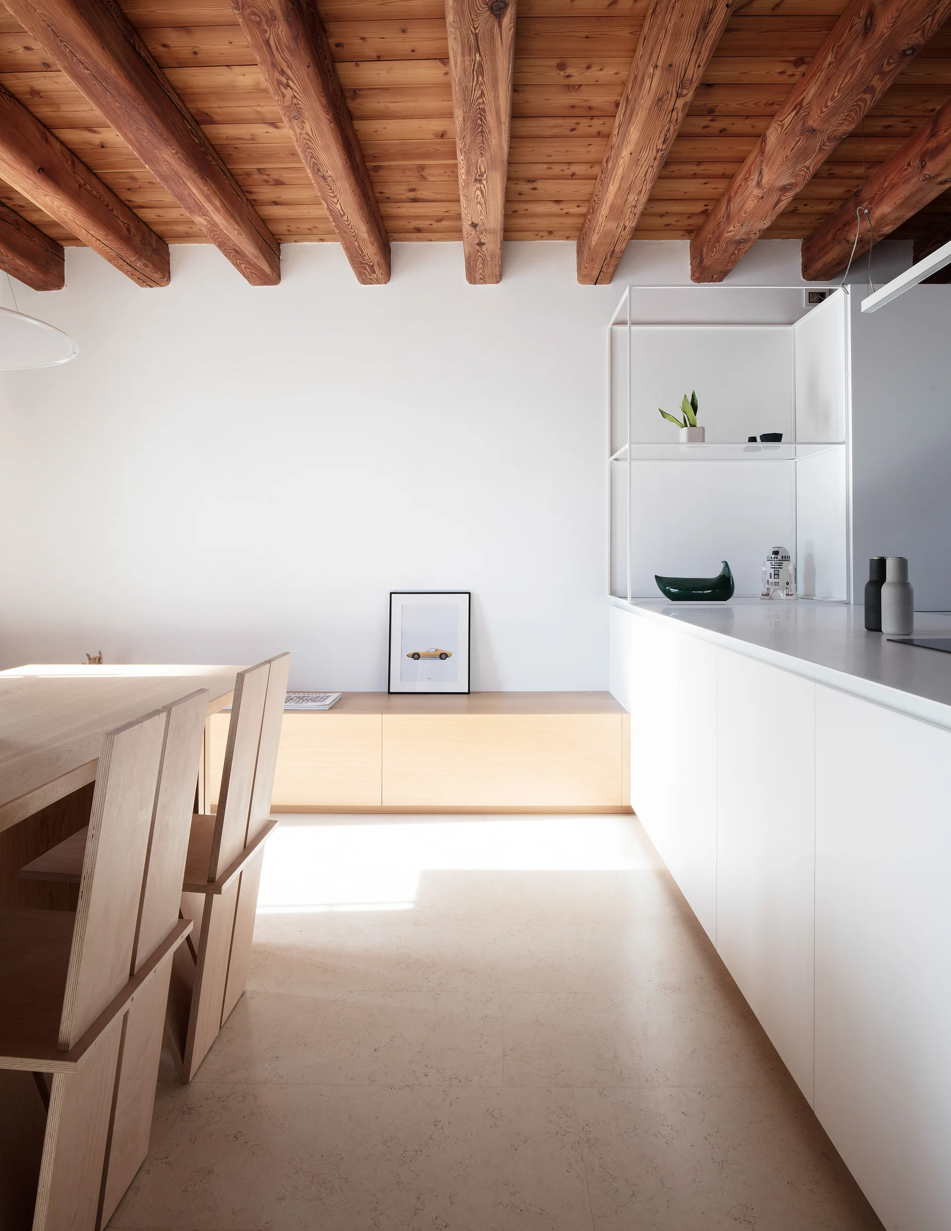 A minimalist kitchen features white flat-front cabinets, a light wood dining set, and an exposed timber ceiling with thick wooden beams.