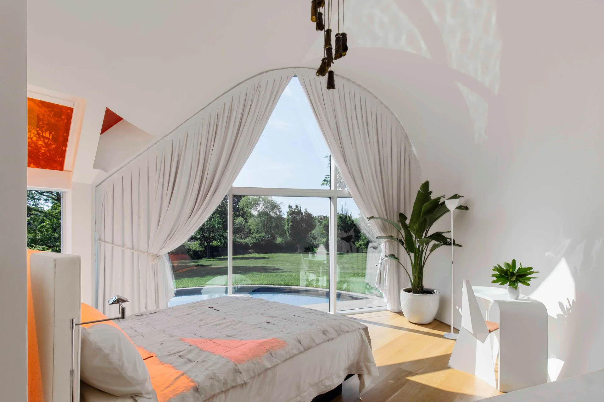 A minimalist bedroom features a bed with neutral linens, a large pointed arched window framed by draped white curtains, and light wood flooring.