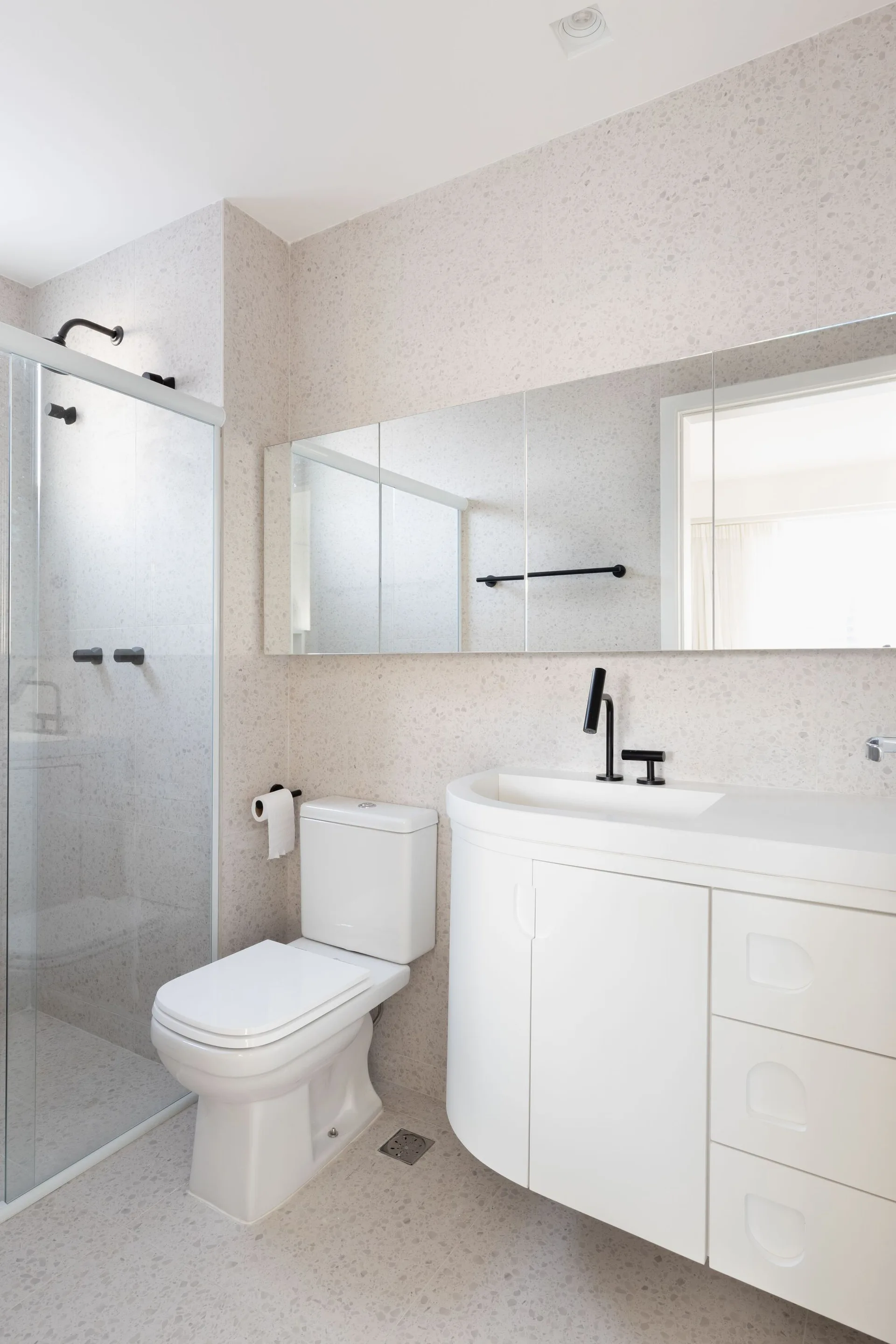 A modern bathroom showcases speckled grey tiles on the floor and walls, a white curved vanity, a glass shower, and matte black fixtures.
