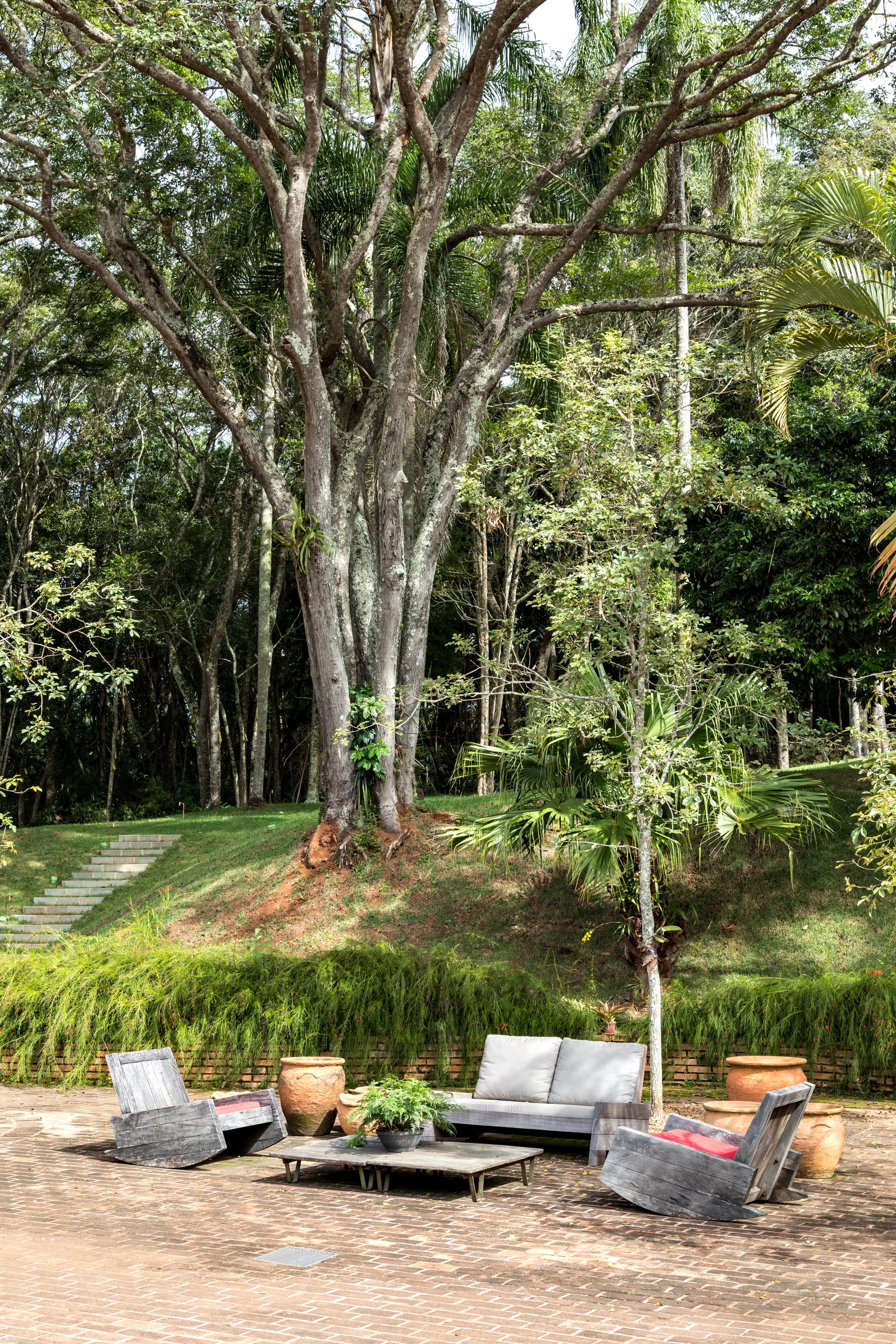 Weathered gray wooden outdoor furniture sits on a brick-paved patio surrounded by large green trees and terracotta planters.