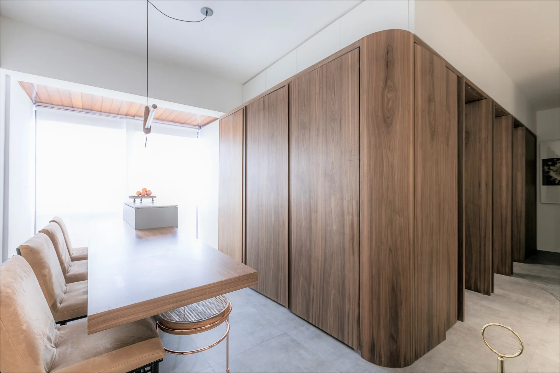 A modern dining room features a long wooden table with upholstered beige chairs alongside a large floor-to-ceiling wood storage wall with a curved corner.