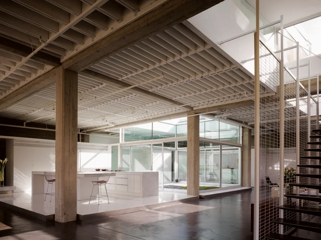 A minimalist white kitchen island stands in a spacious loft featuring exposed ceiling joists, concrete pillars, and a glass-enclosed atrium.