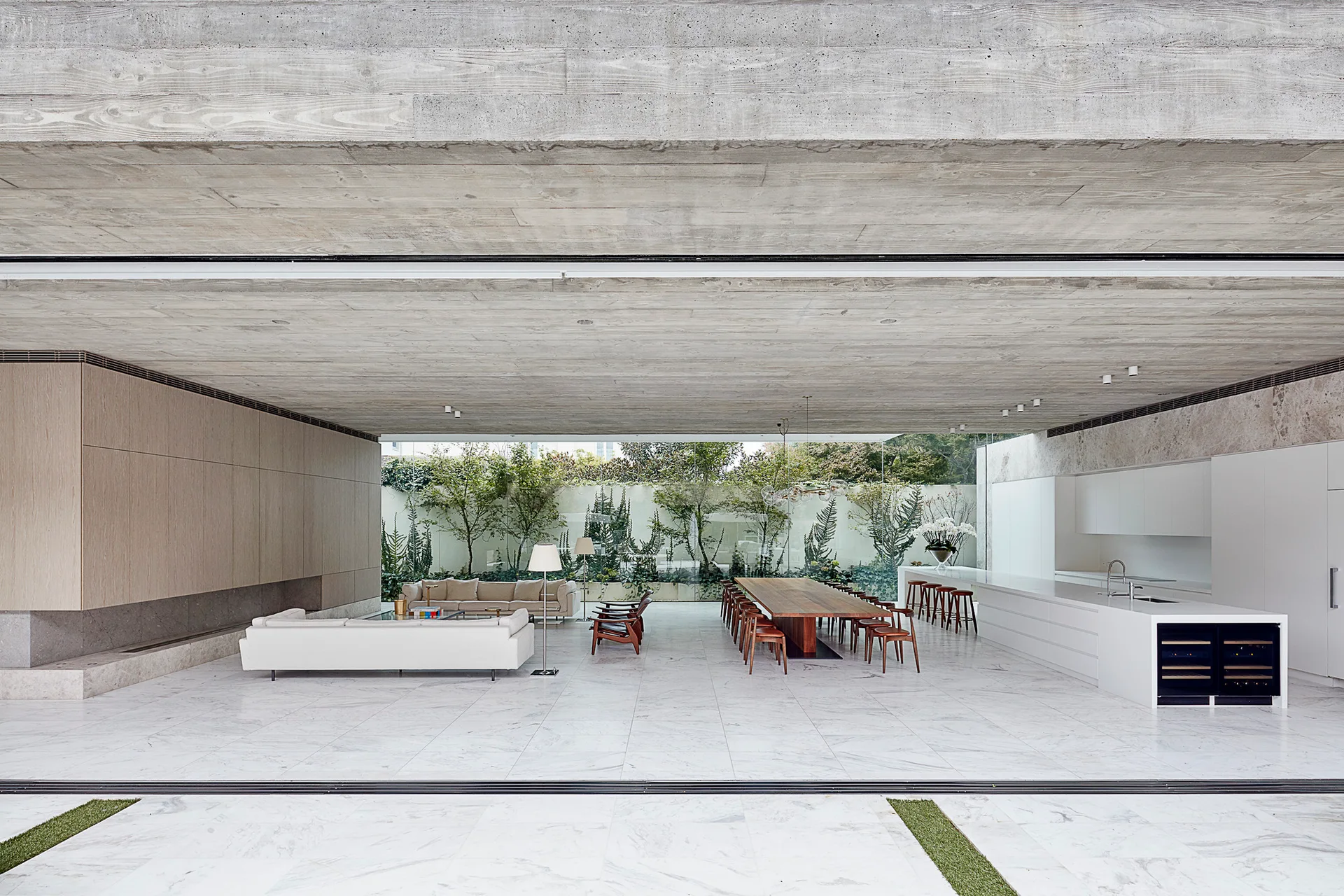 A modern open-concept living area features a textured concrete ceiling, white marble tile flooring, a long wood dining table, and kitchen island.