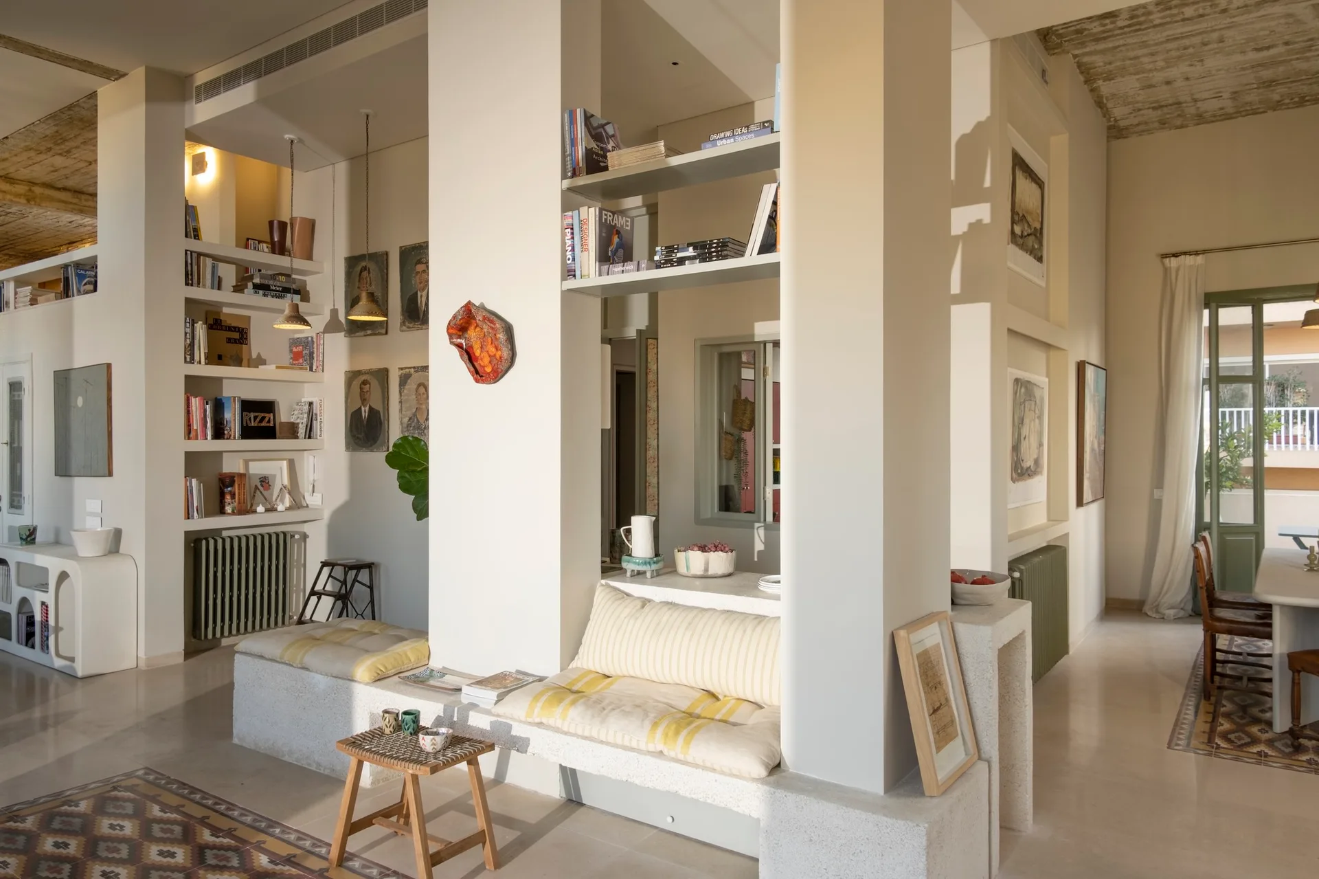 An open-plan room featuring thick plaster columns with built-in wooden shelves and a stone-like bench topped with yellow striped cushions.