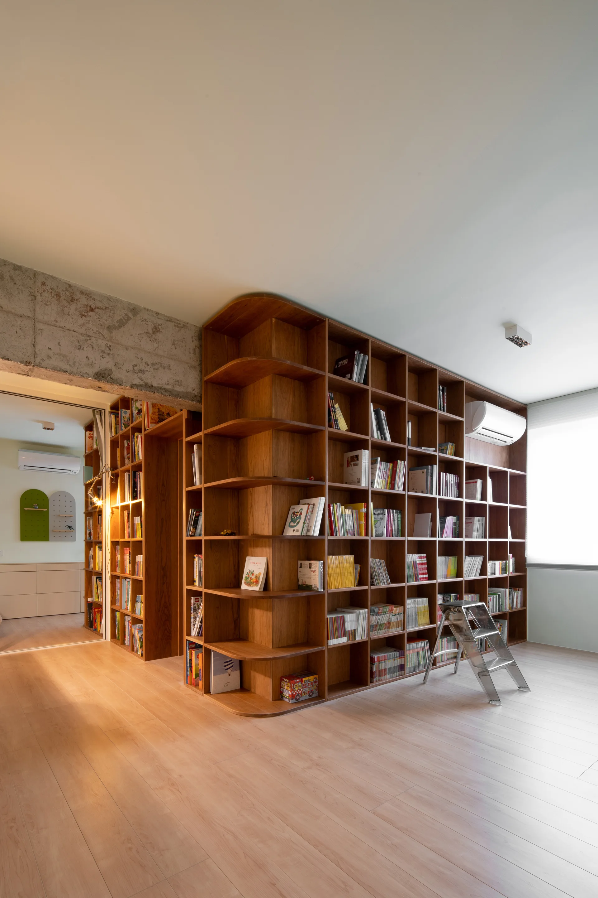 A large wooden built-in bookshelf with a curved corner stands next to an open doorway under an exposed concrete beam.