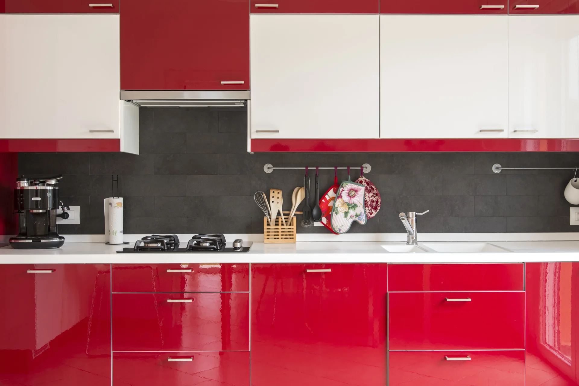 A modern kitchen with glossy red lower cabinets, white upper cabinets, a dark grey tiled backsplash, and a white countertop with a gas stovetop.