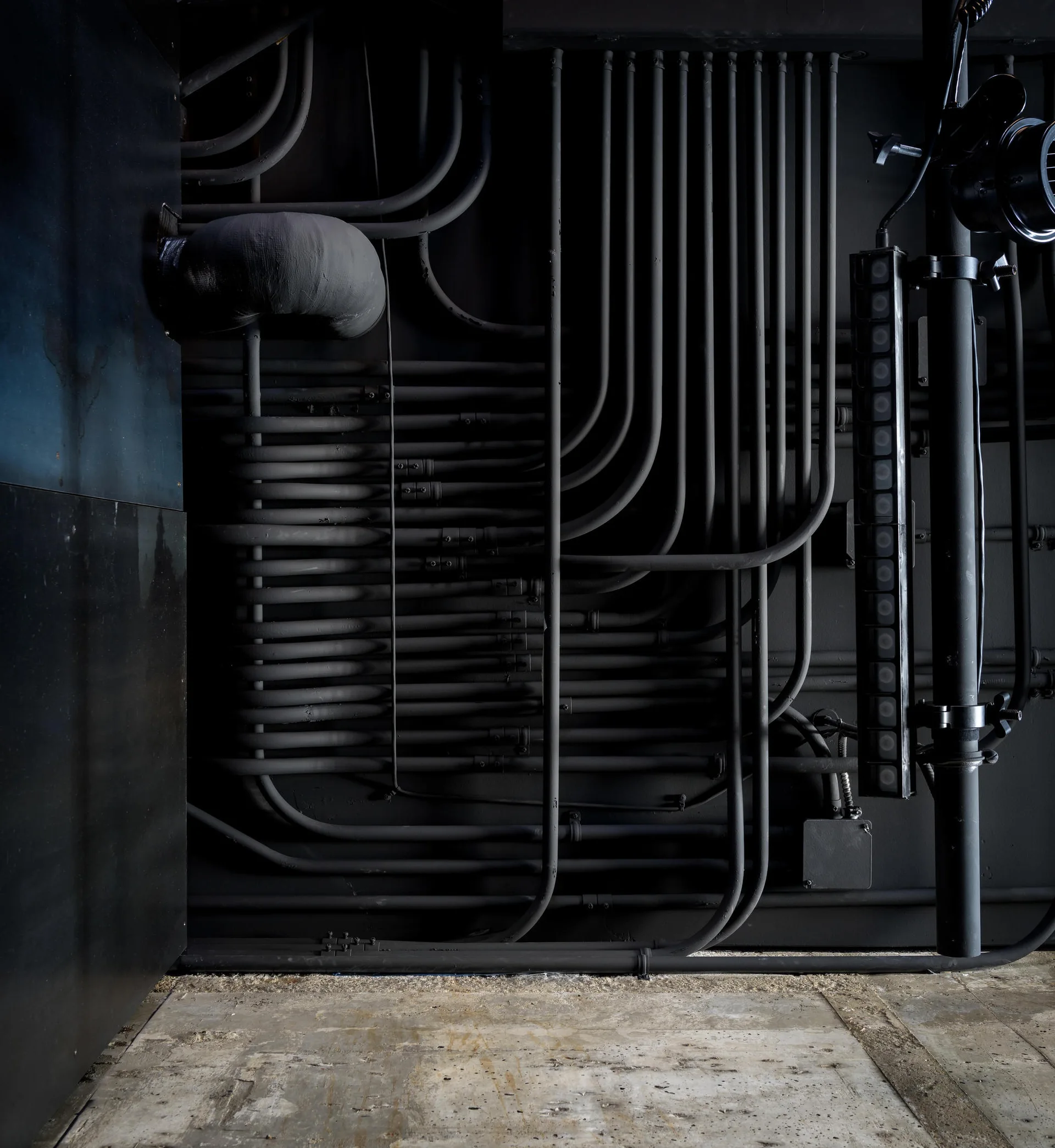 Numerous black painted conduits and utility pipes run vertically and horizontally across a matte black wall above an unfinished concrete floor.