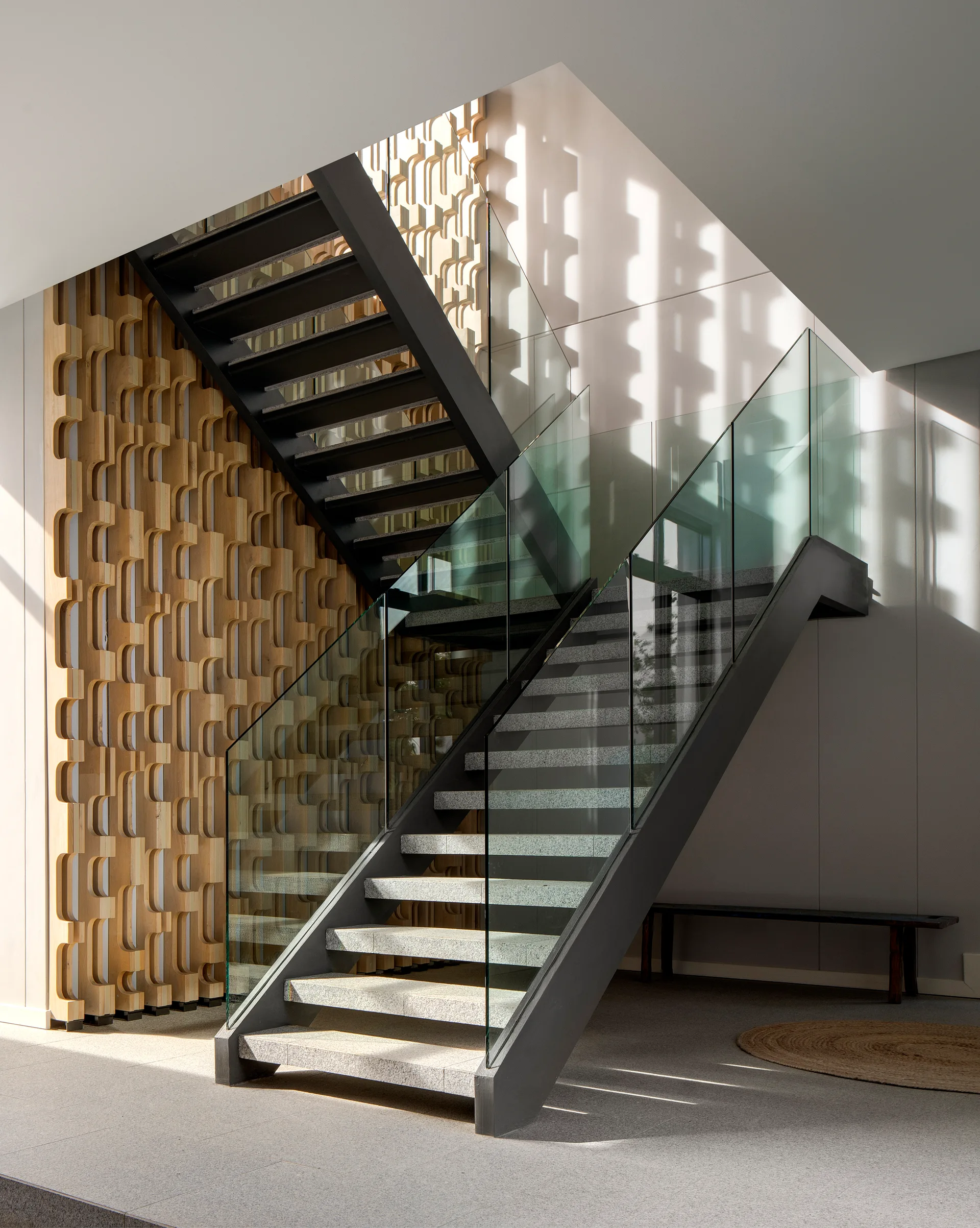 A modern steel and glass staircase rises in front of a light wood wall with a sculptural geometric pattern, with sunlight casting shadows.