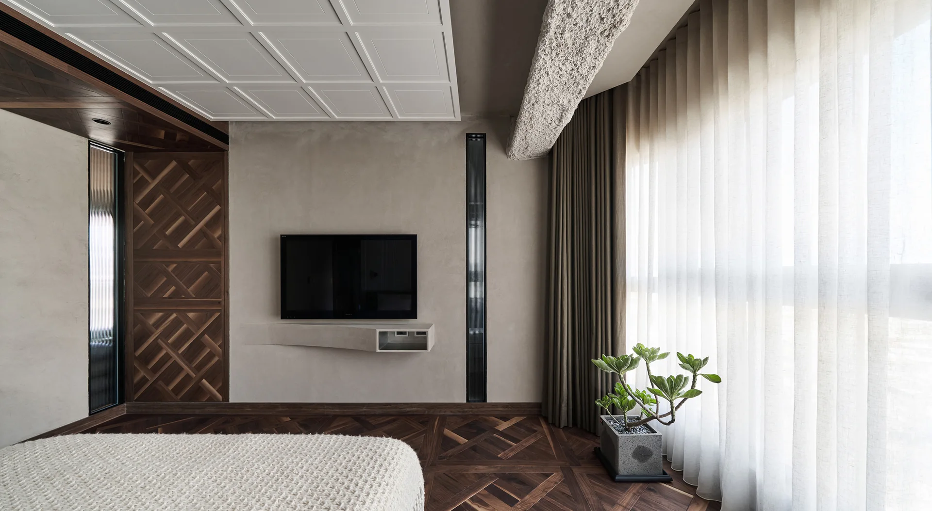 A modern bedroom features a wall-mounted TV with a plaster floating shelf, intricate geometric parquet wood floors, and floor-to-ceiling sheer curtains.