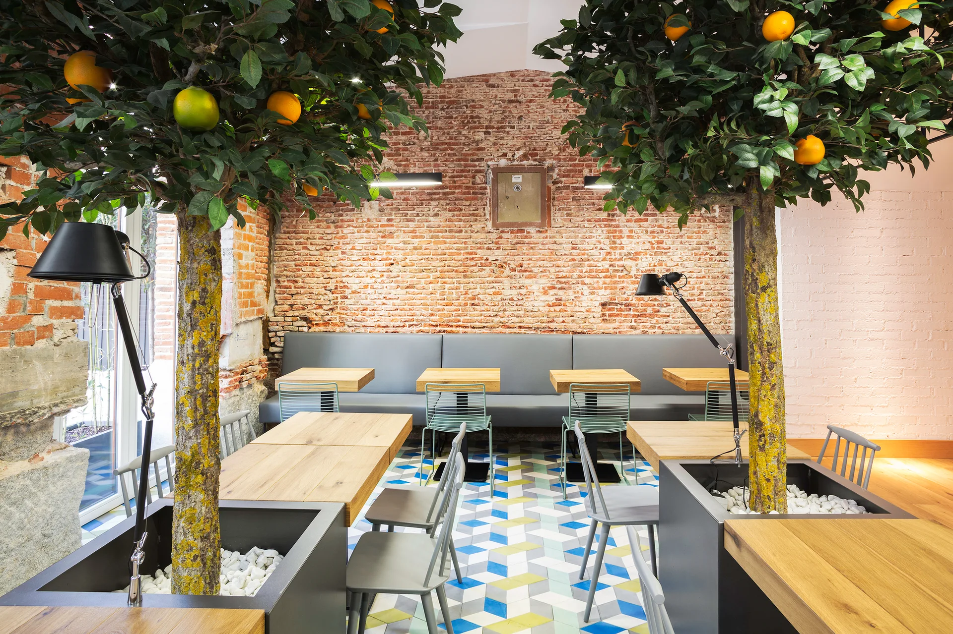 A modern dining area featuring an exposed brick wall, geometric cube patterned flooring, grey banquette seating, and large planters with indoor citrus trees.