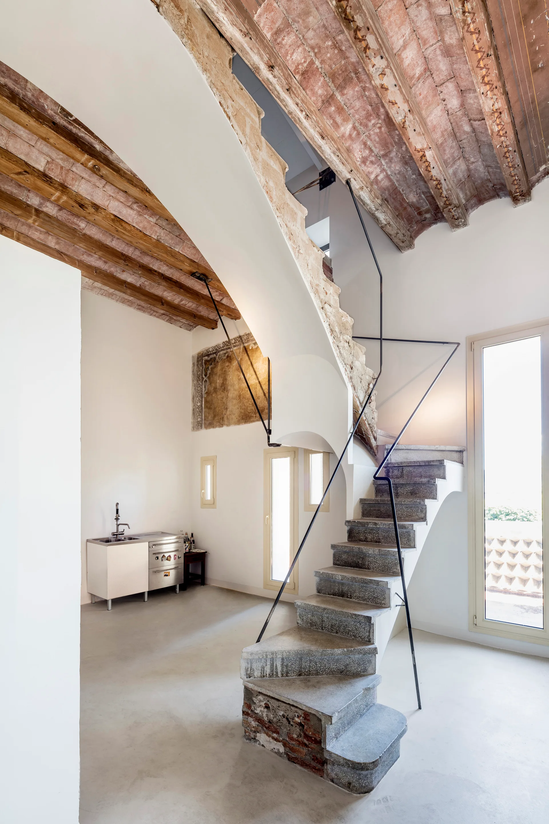 A sculptural stone staircase with a thin black metal railing stands under an exposed vaulted brick ceiling in a white minimalist room.