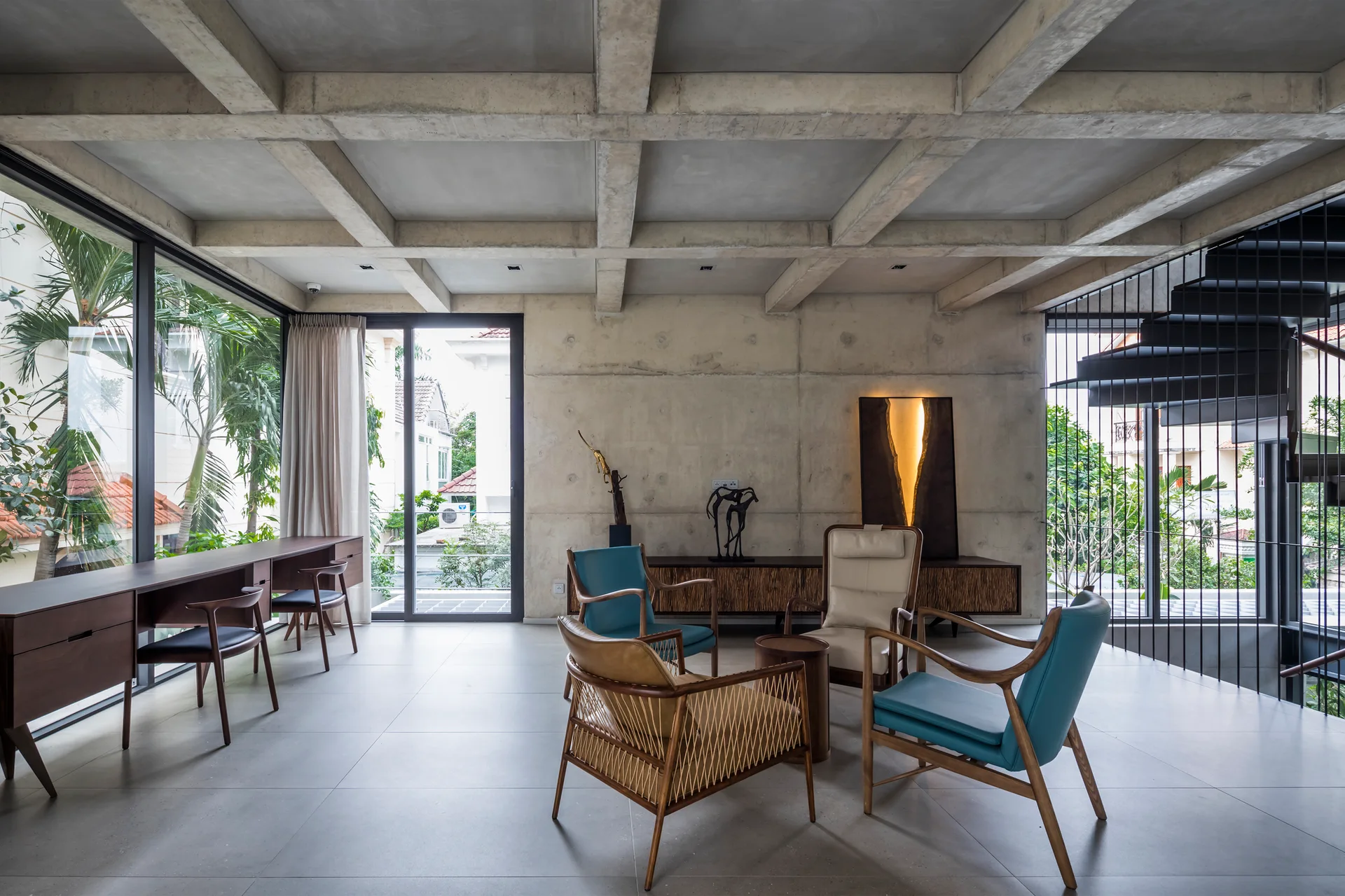 A spacious concrete living room features a grid ceiling, a cluster of mid-century armchairs, a long desk by the windows, and a black spiral staircase.