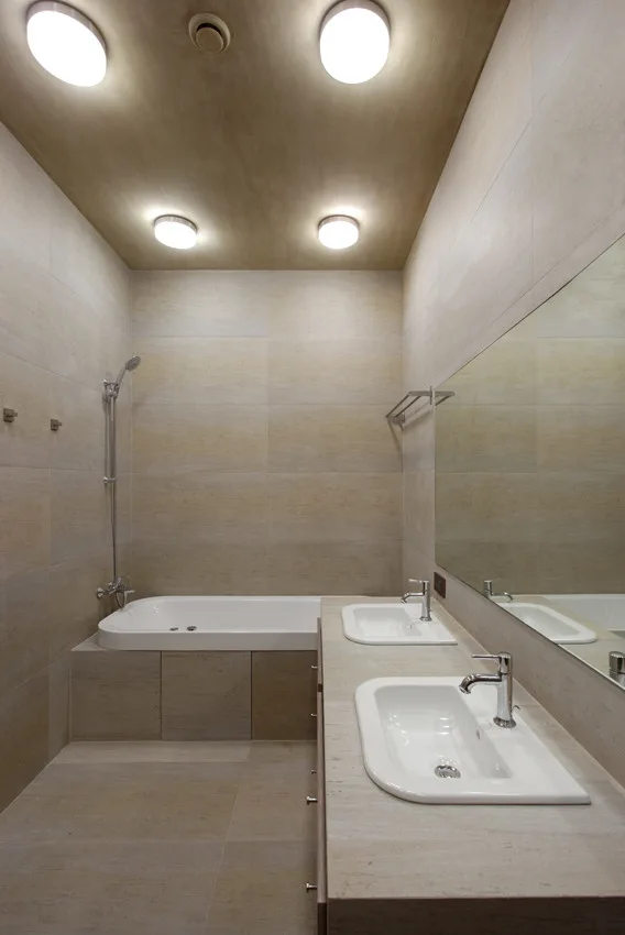 A modern beige tiled bathroom features a long stone-like double vanity with white sinks, a matching bathtub, and round ceiling lights.