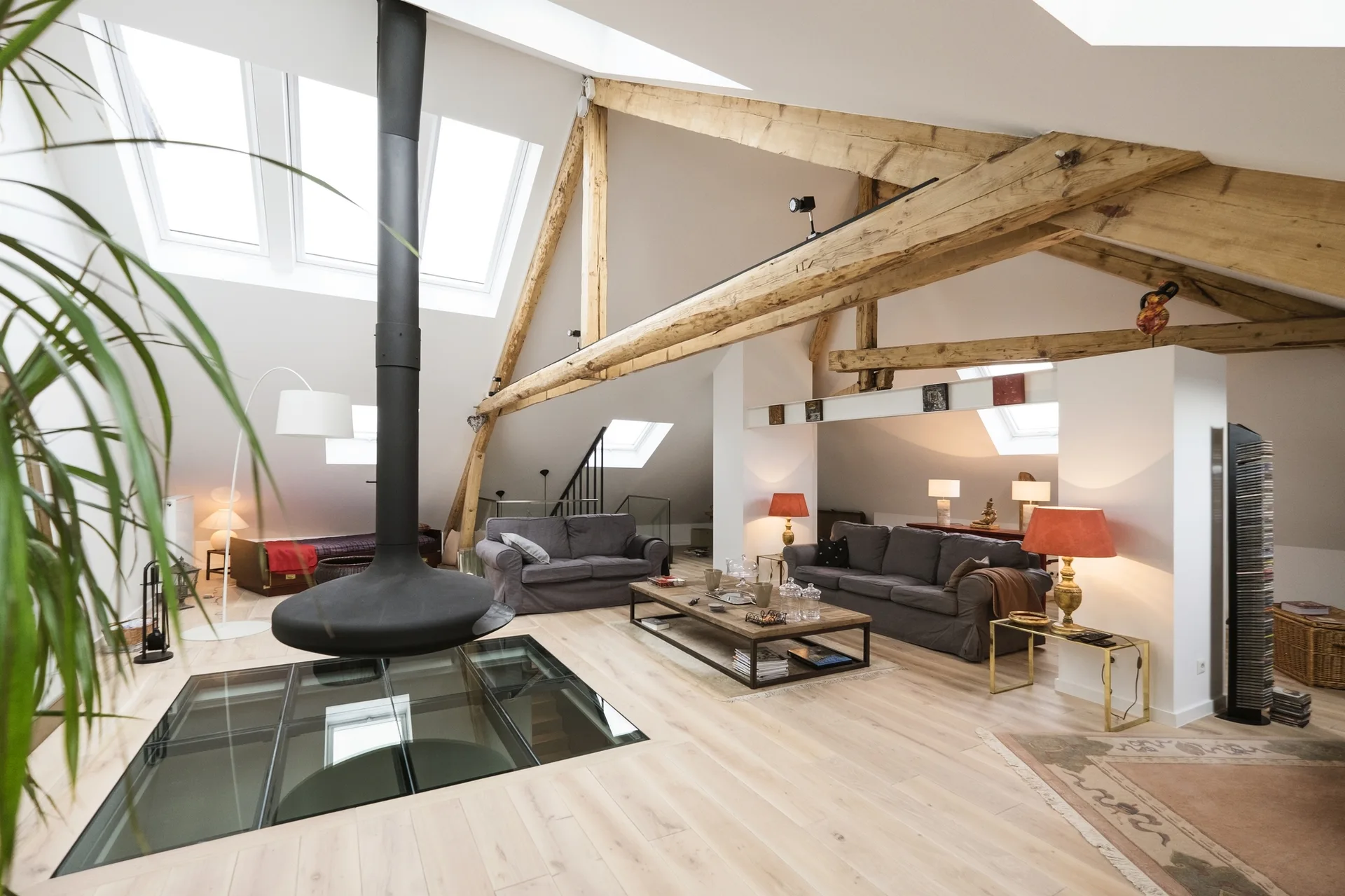 A spacious attic living room with exposed wood beams, skylights, two gray sofas, a suspended black fireplace, and a glass floor section.