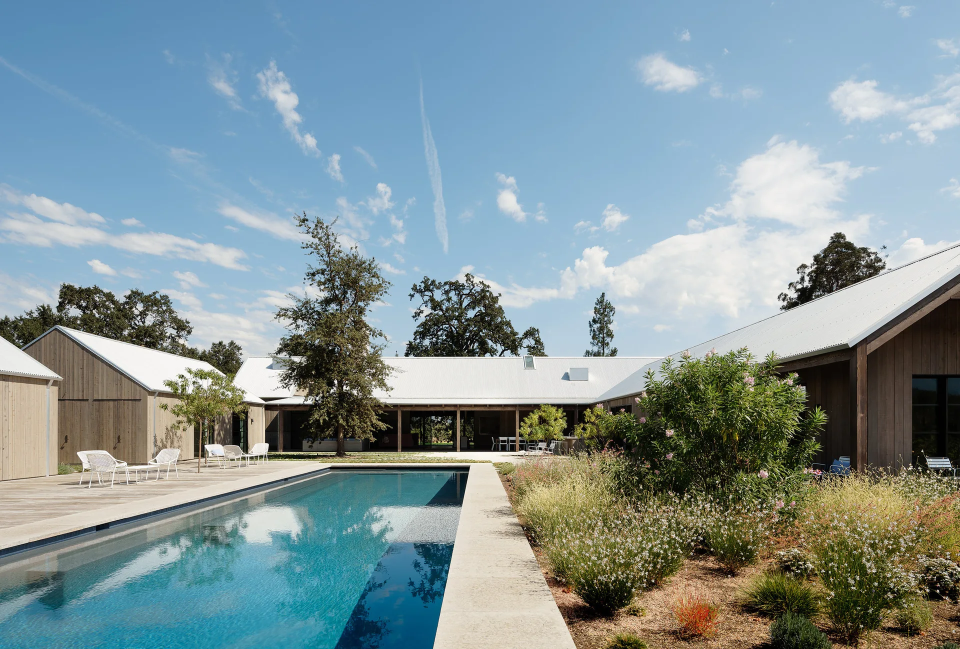 A long rectangular swimming pool sits in a courtyard surrounded by weathered wood-clad buildings with white gabled roofs and natural landscaping.