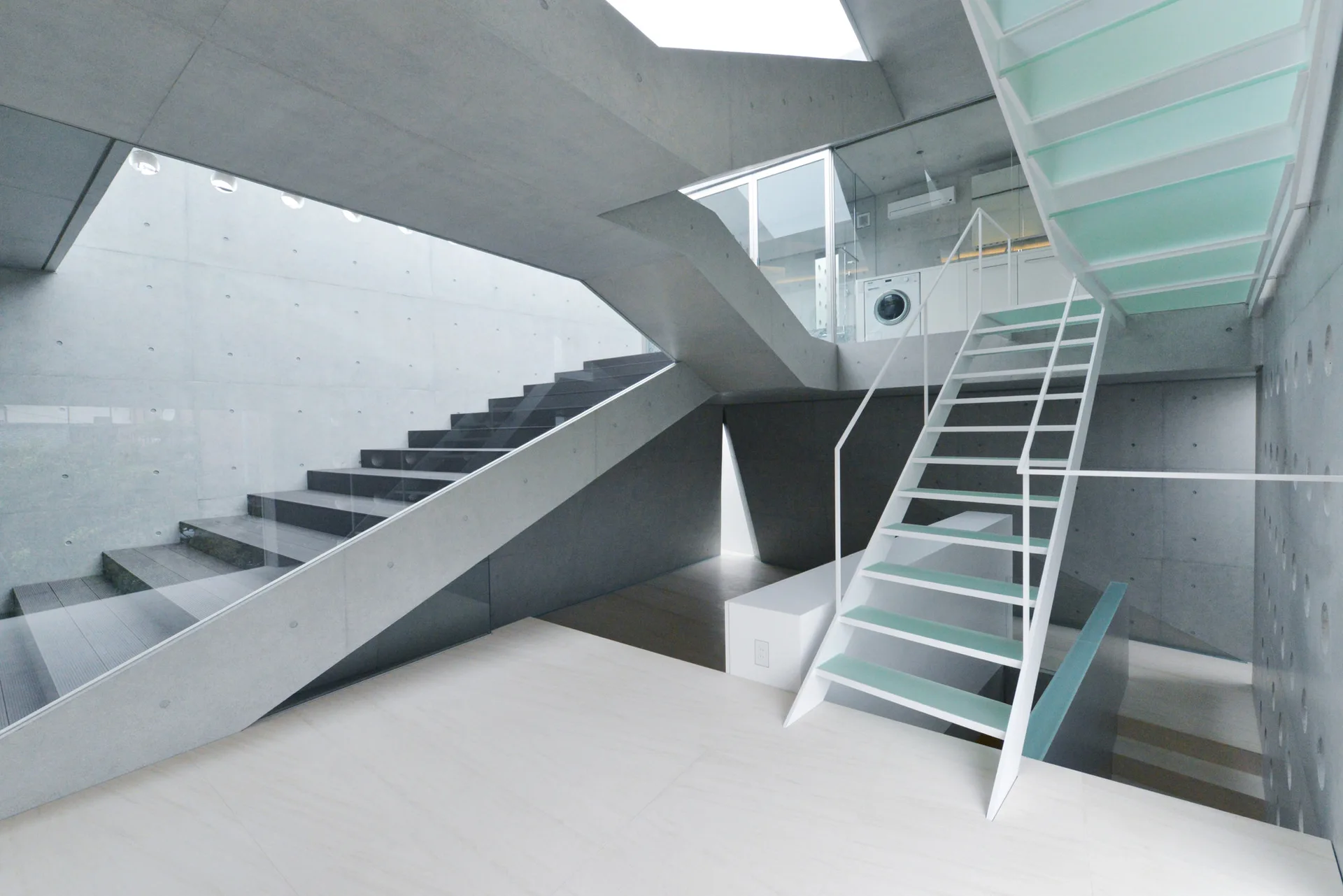 A minimalist grey concrete stairwell features an open-tread white metal staircase with mint green steps alongside a dark concrete staircase.
