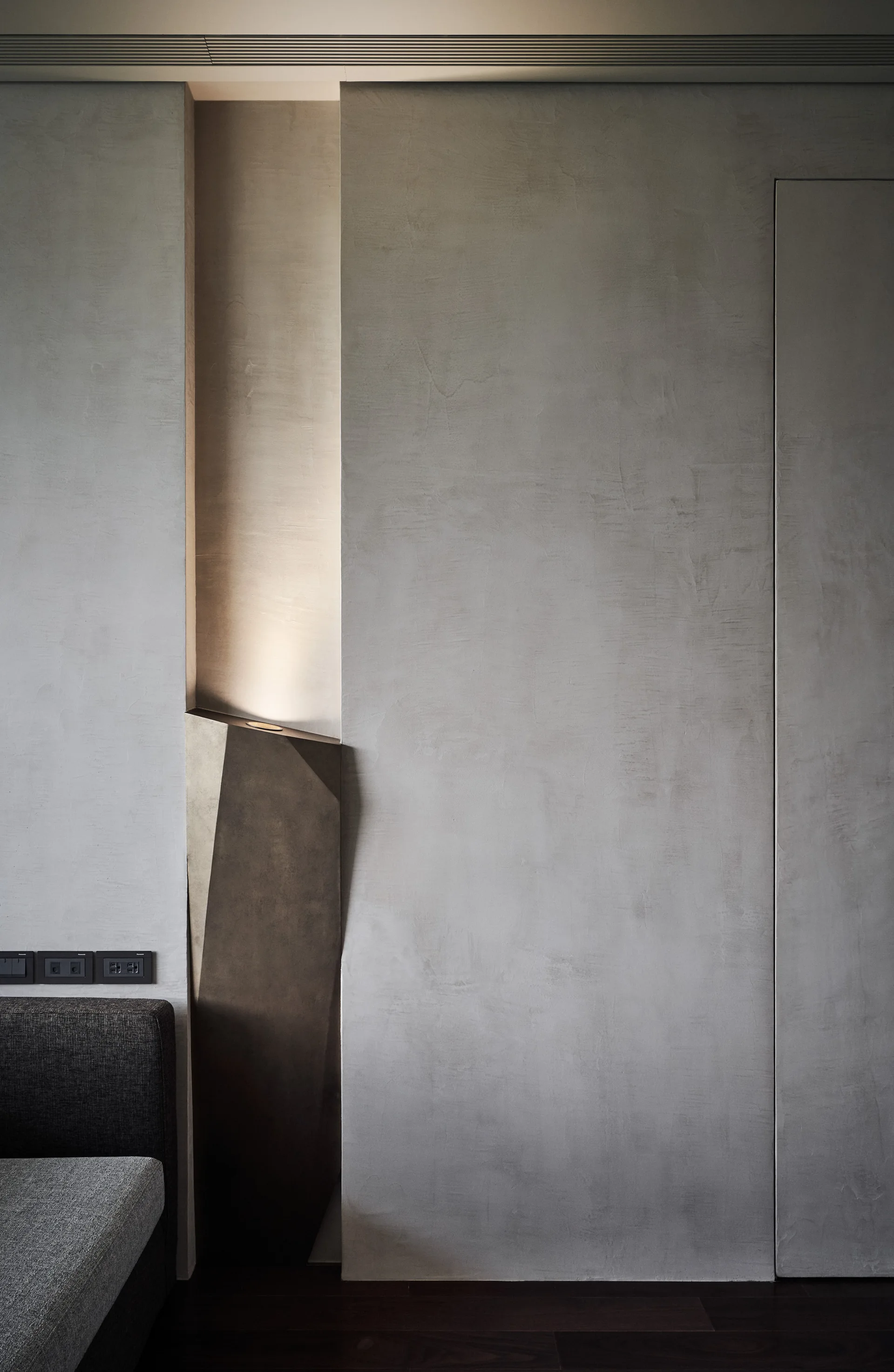 A vertical recess in a grey textured plaster wall houses an angular sculptural light casting a warm glow next to a grey fabric sofa.