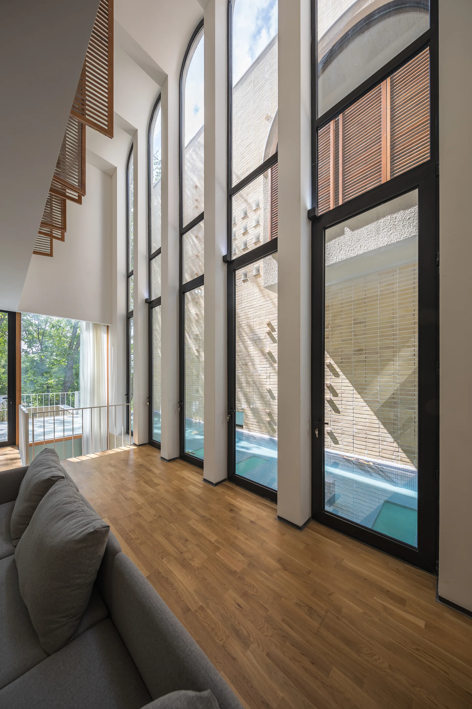 A modern living space with a grey sofa, hardwood floors, and tall black-framed arched windows overlooking an outdoor pool.
