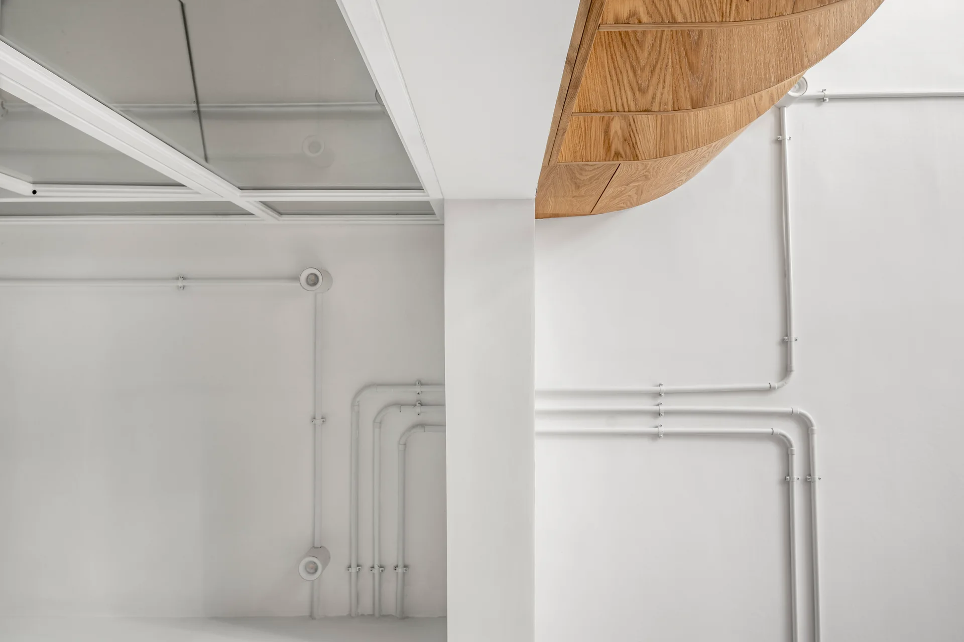 An interior detail showing a clean white wall with matching painted conduits beneath a curved wood paneled ceiling and a glass skylight grid.
