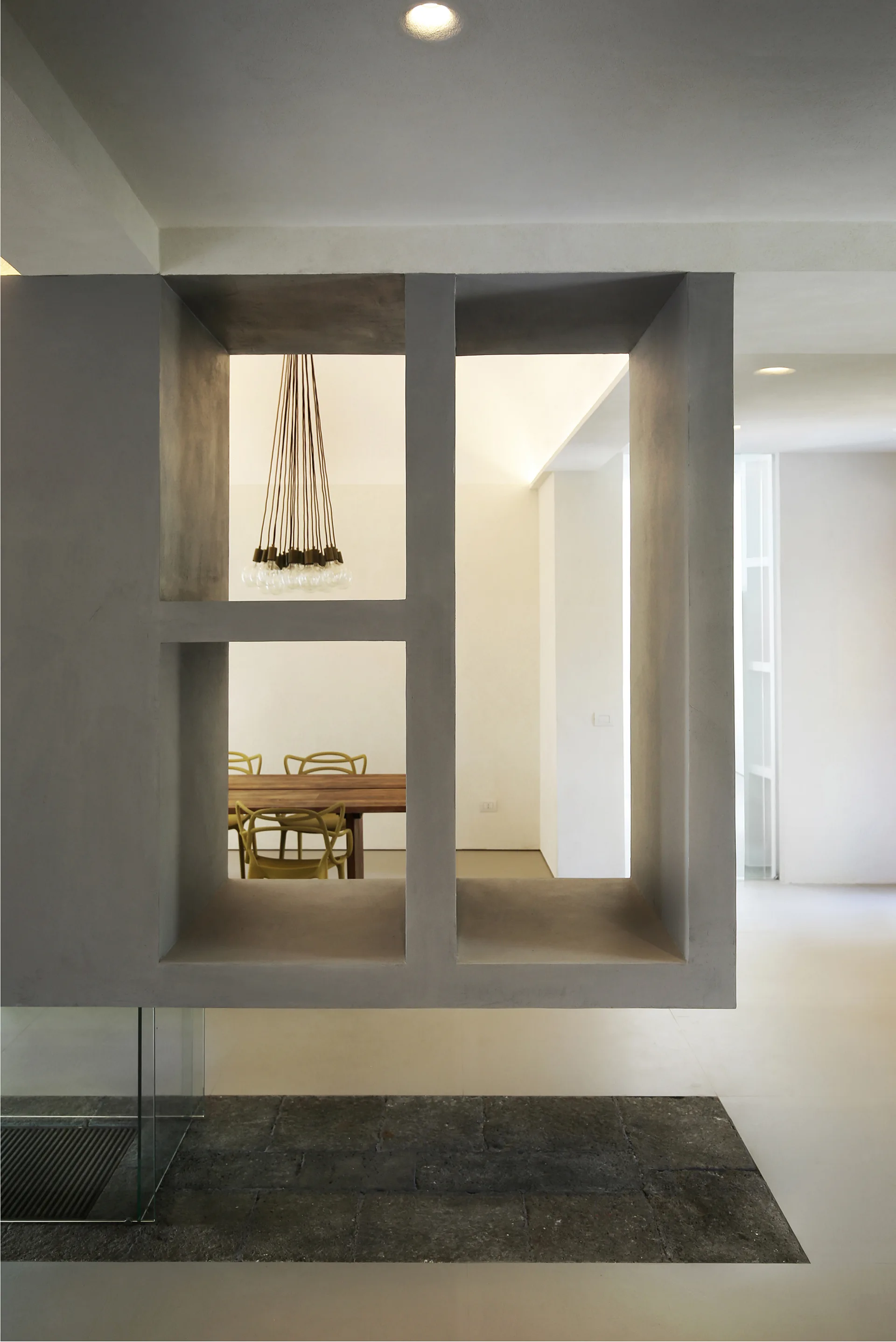 A grey concrete partition wall with rectangular openings frames a dining room with a wooden table, yellow chairs, and a bulb pendant light.