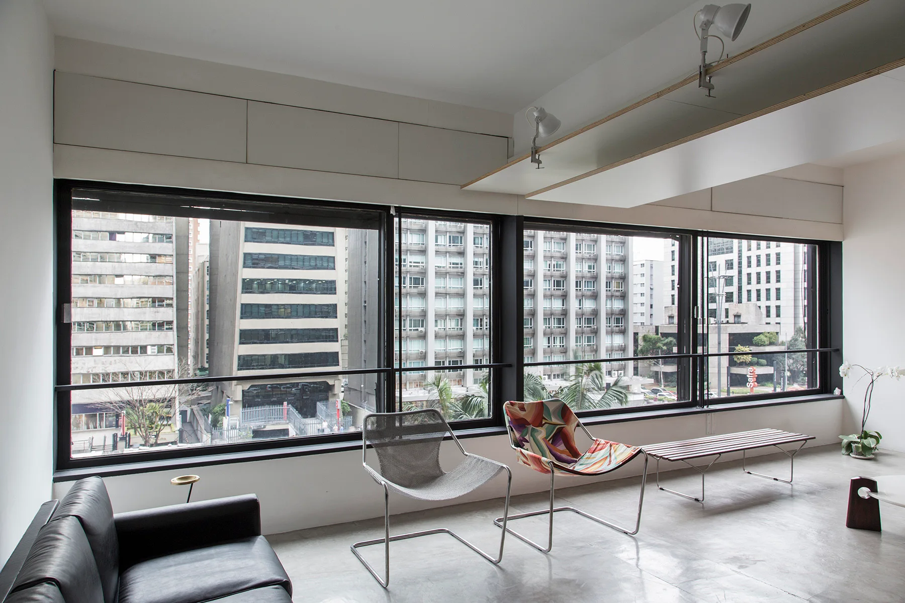 An industrial loft living room features concrete flooring, modern sling chairs, and a slatted bench facing large windows with a cityscape view.