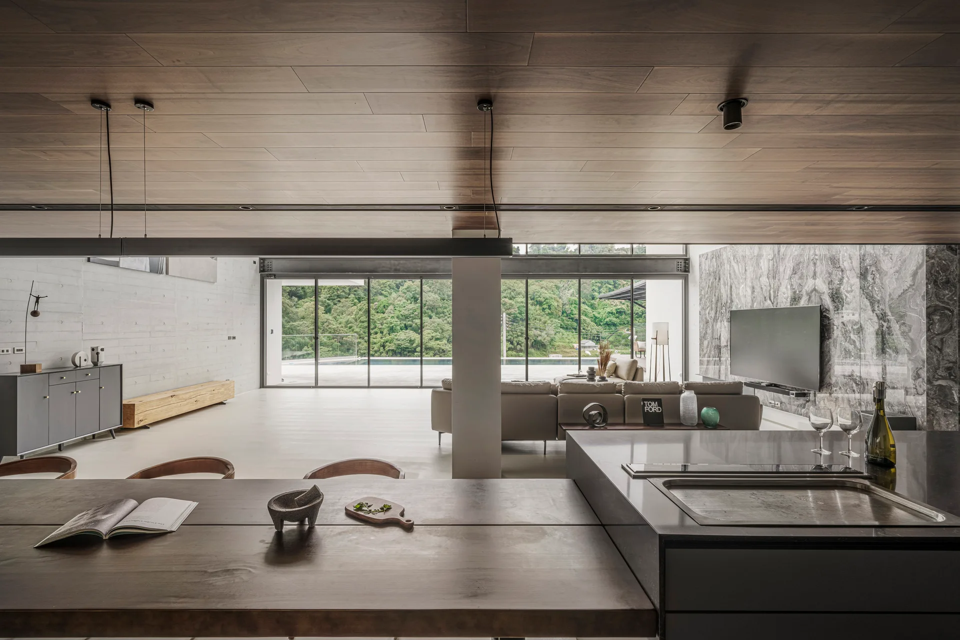 An open-concept home interior featuring a wood-plank ceiling, kitchen island, and a distant living room with sliding glass doors opening to greenery.