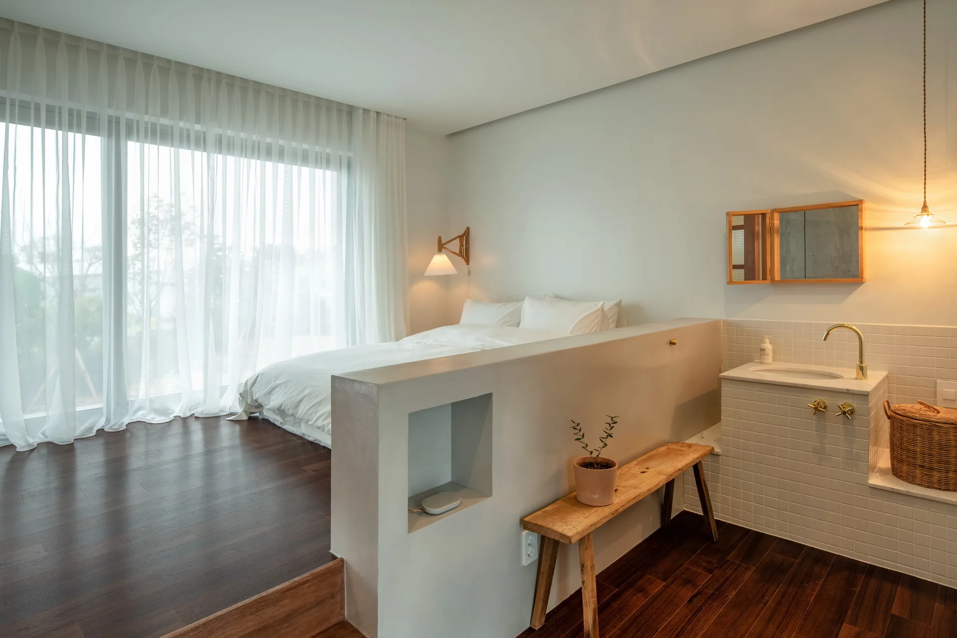 A minimalist bedroom features a low partition wall dividing a platform bed with white bedding from a small tiled sink vanity on dark wood floors.
